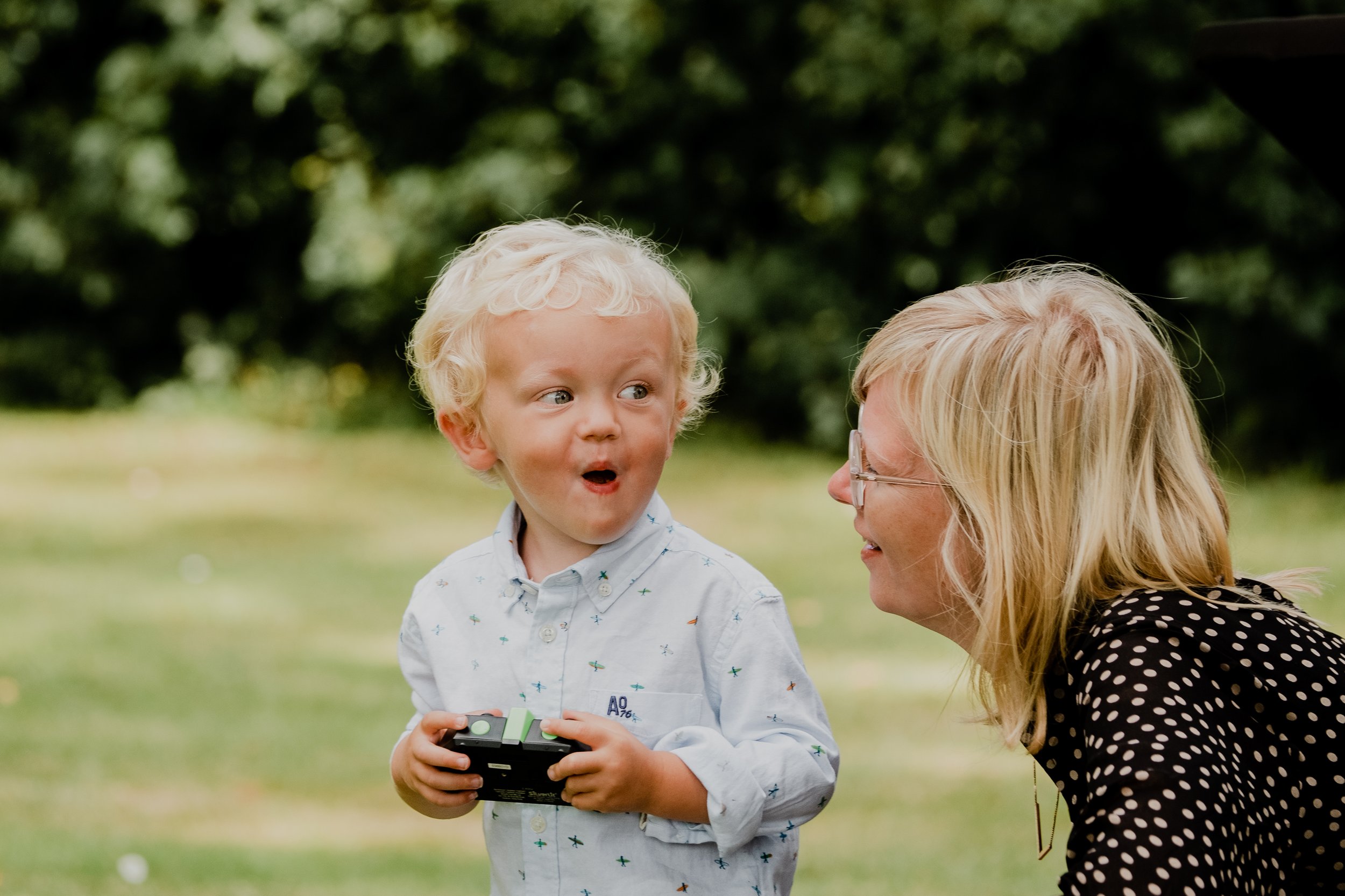 A young boy with curly blond hair holding a small toy or device, looking at a woman with glasses and blond hair, both outdoors with blurred greenery in the background.