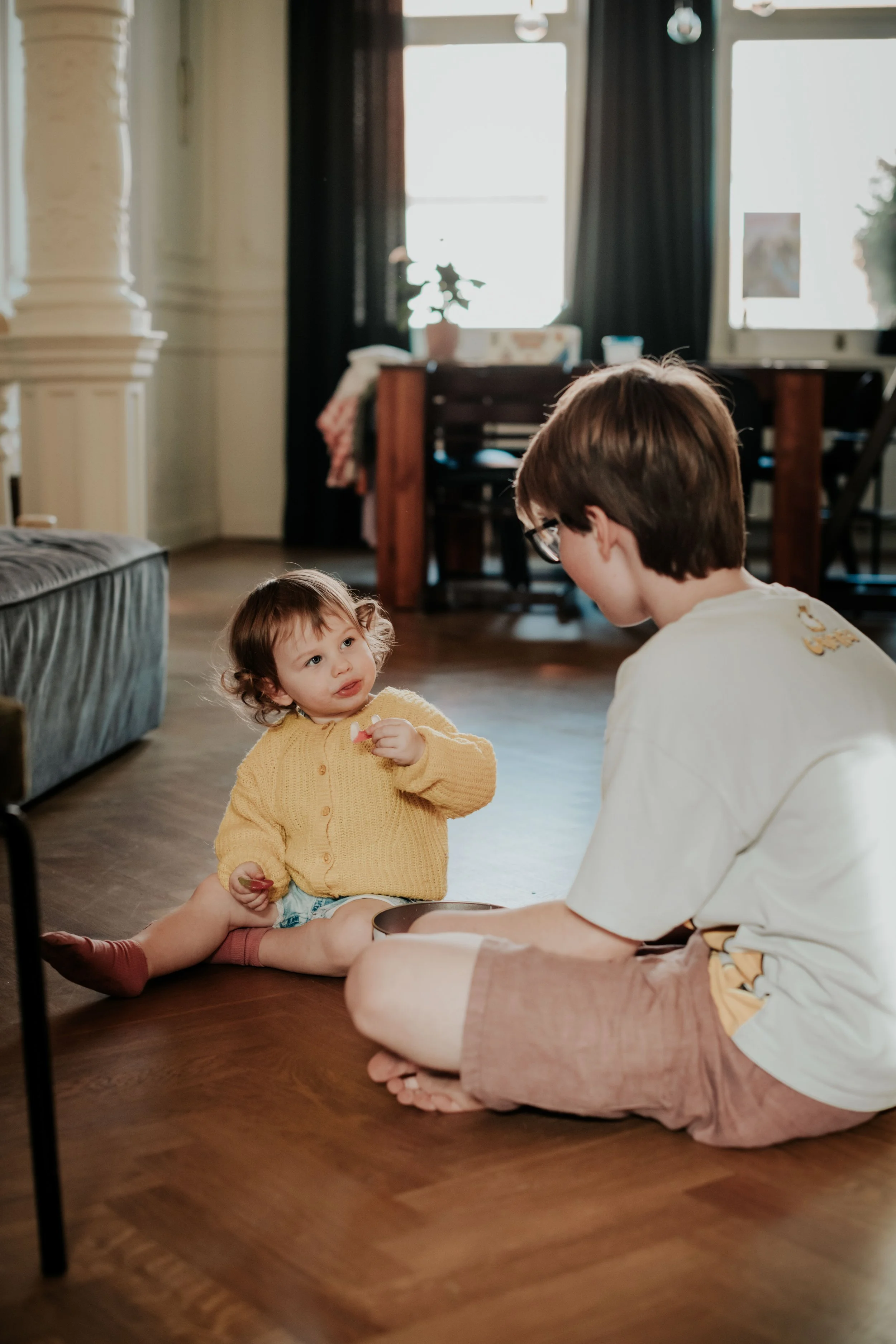 A young woman with short brown hair and glasses sitting cross-legged on a wooden floor, talking to a small girl with curly hair wearing a yellow sweater. They are in a cozy living room with dark curtains, a table, and a sofa in the background.