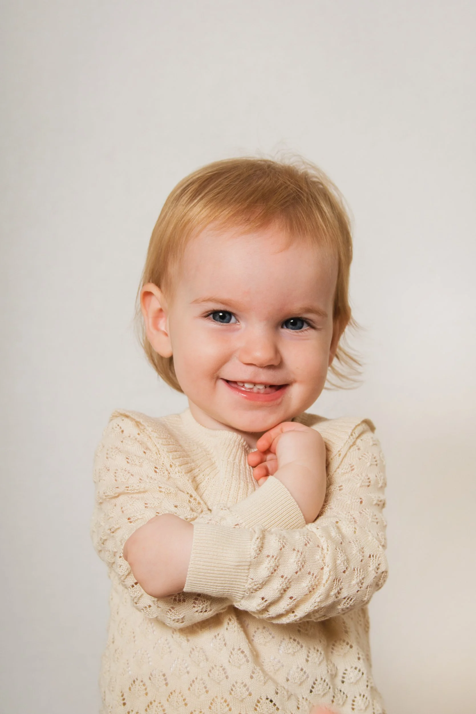 A young child with red hair and blue eyes smiling and holding their chin with one hand, wearing a cream-colored, lacy sweater.