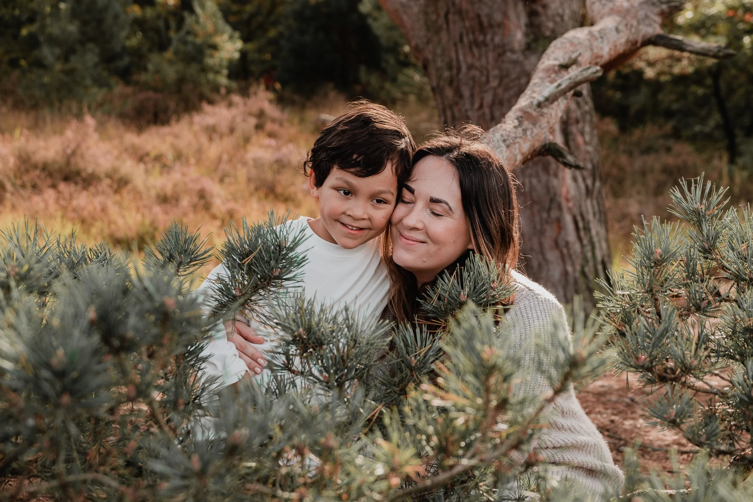 A woman and a young boy embracing in a forested area with trees and bushes around them.
