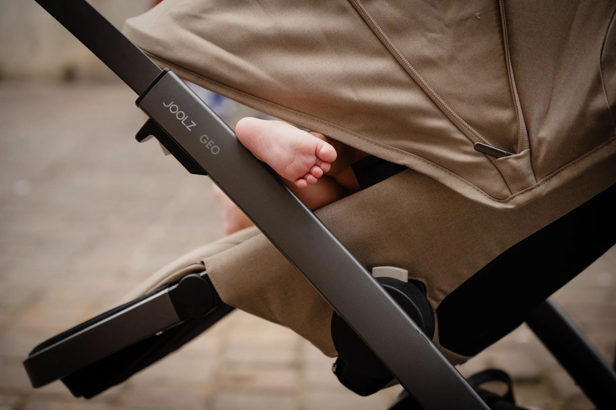 Close-up of a baby’s hand gripping the metal frame of a stroller, with beige fabric and a zipper visible on the stroller
