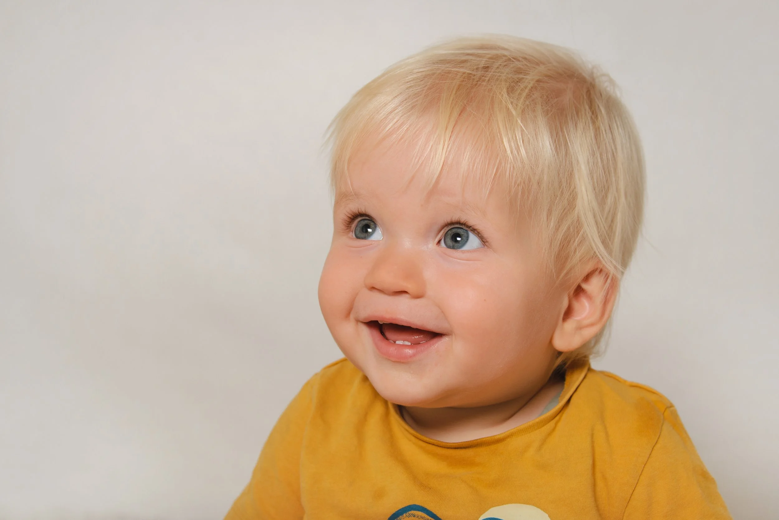 A young child with blonde hair and blue eyes smiling and looking upward, wearing a yellow shirt.