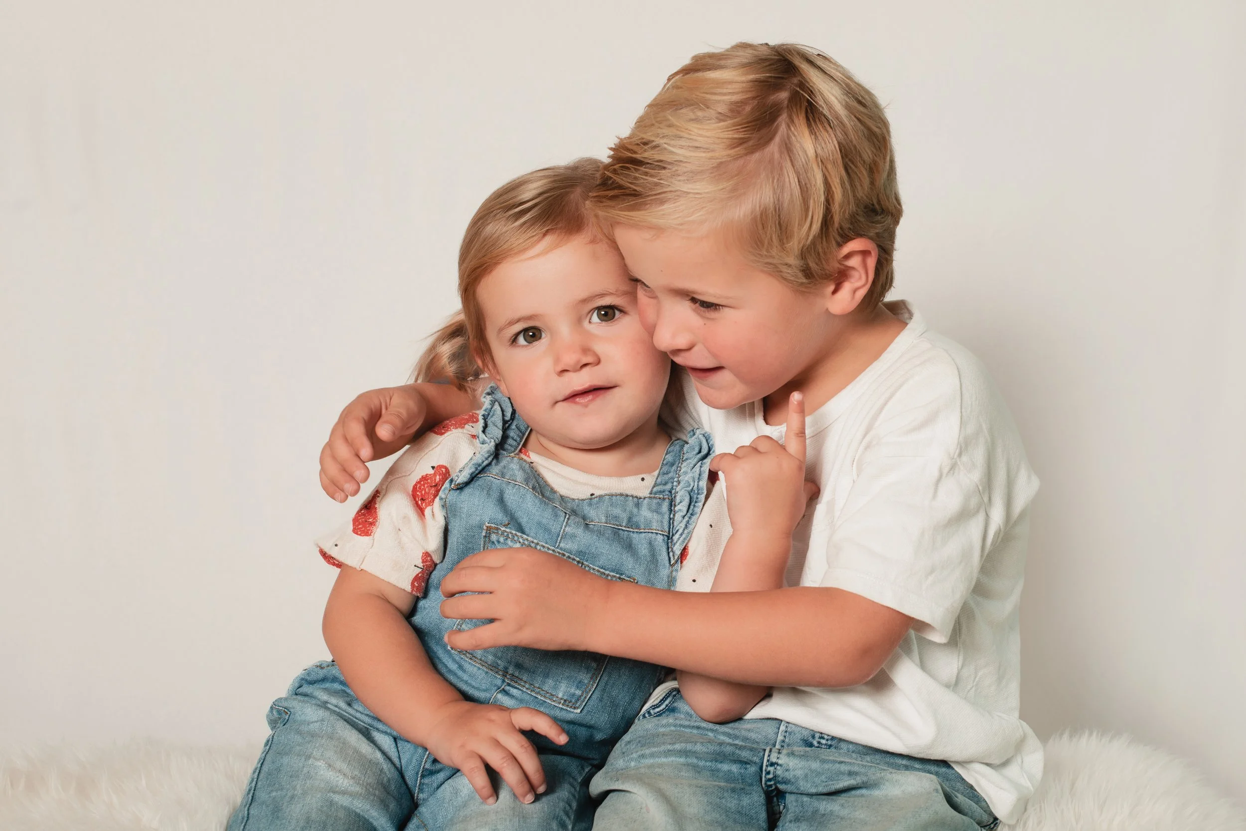 Two children, a girl and a boy, sitting close together against a plain background, with the boy hugging the girl. The girl has light hair and is wearing a white shirt with a floral pattern and denim overalls. The boy has blonde hair and is wearing a 