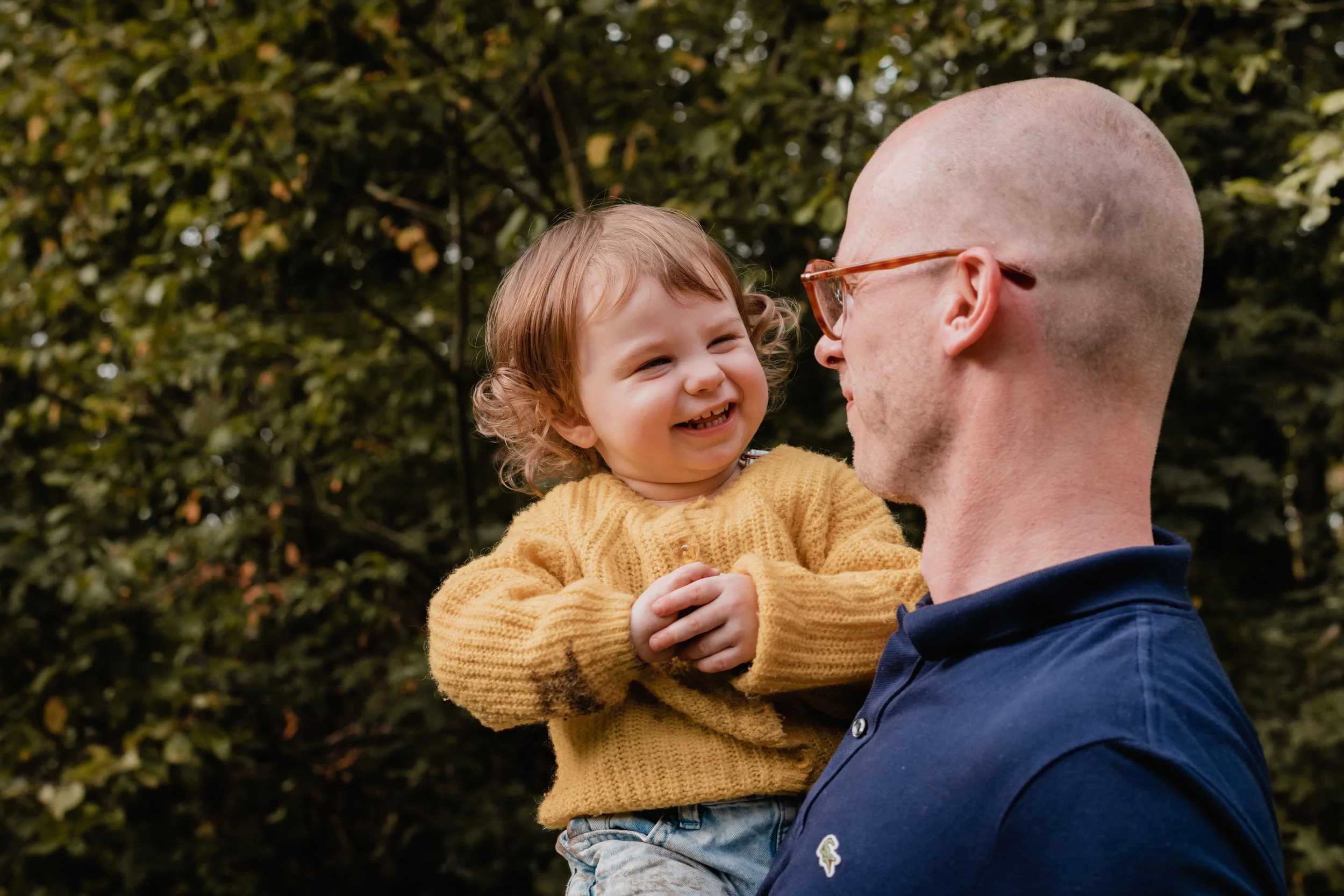 A young child with curly hair smiling and laughing while being held by a man with glasses and a shaved head outdoors with trees in the background.