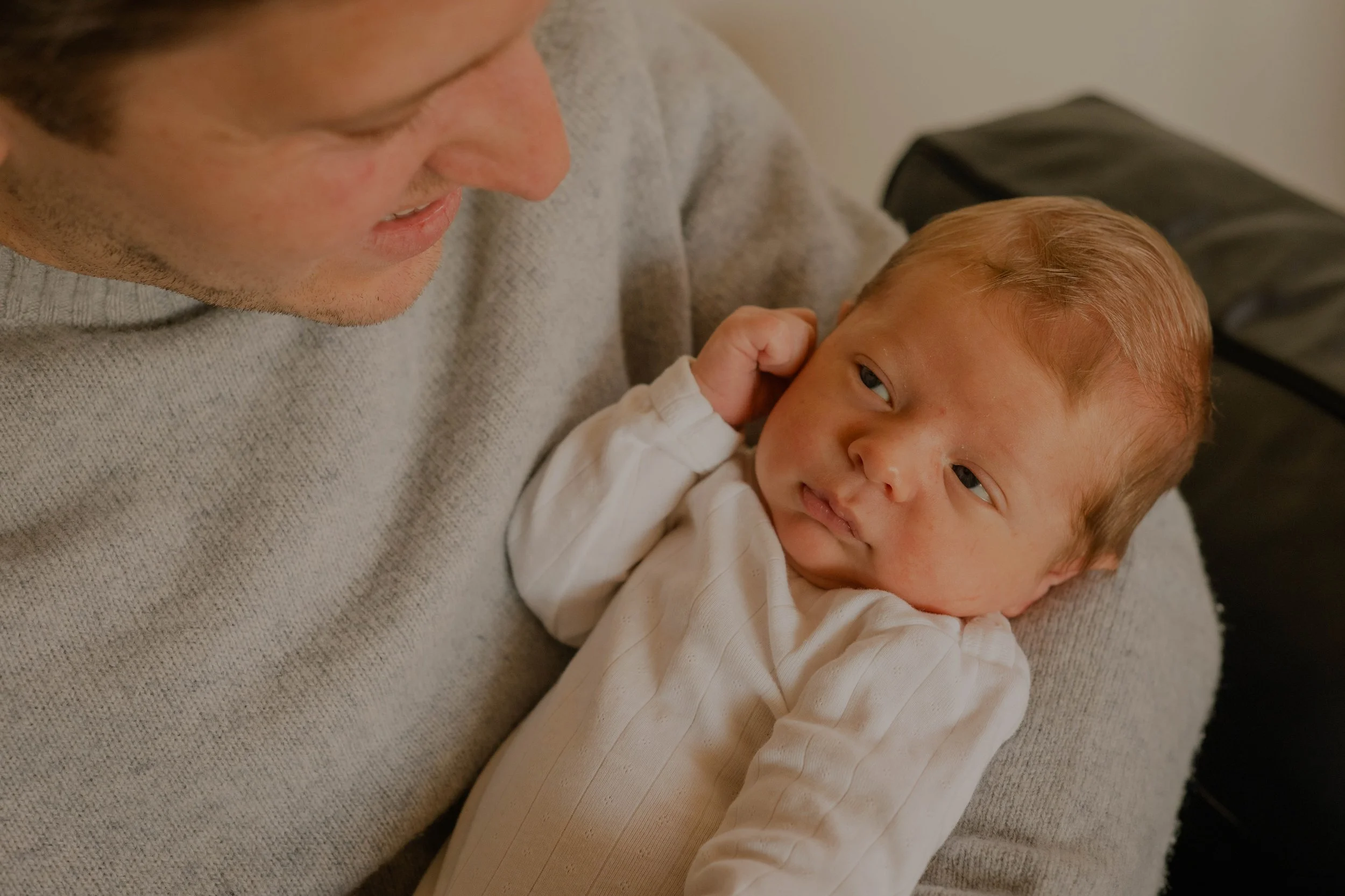 A man with short brown hair and a light beard, wearing a gray sweater, is holding and looking at a young child with blonde hair and a light complexion. The child is lying on the man's lap, looking up with a curious expression. The background includes