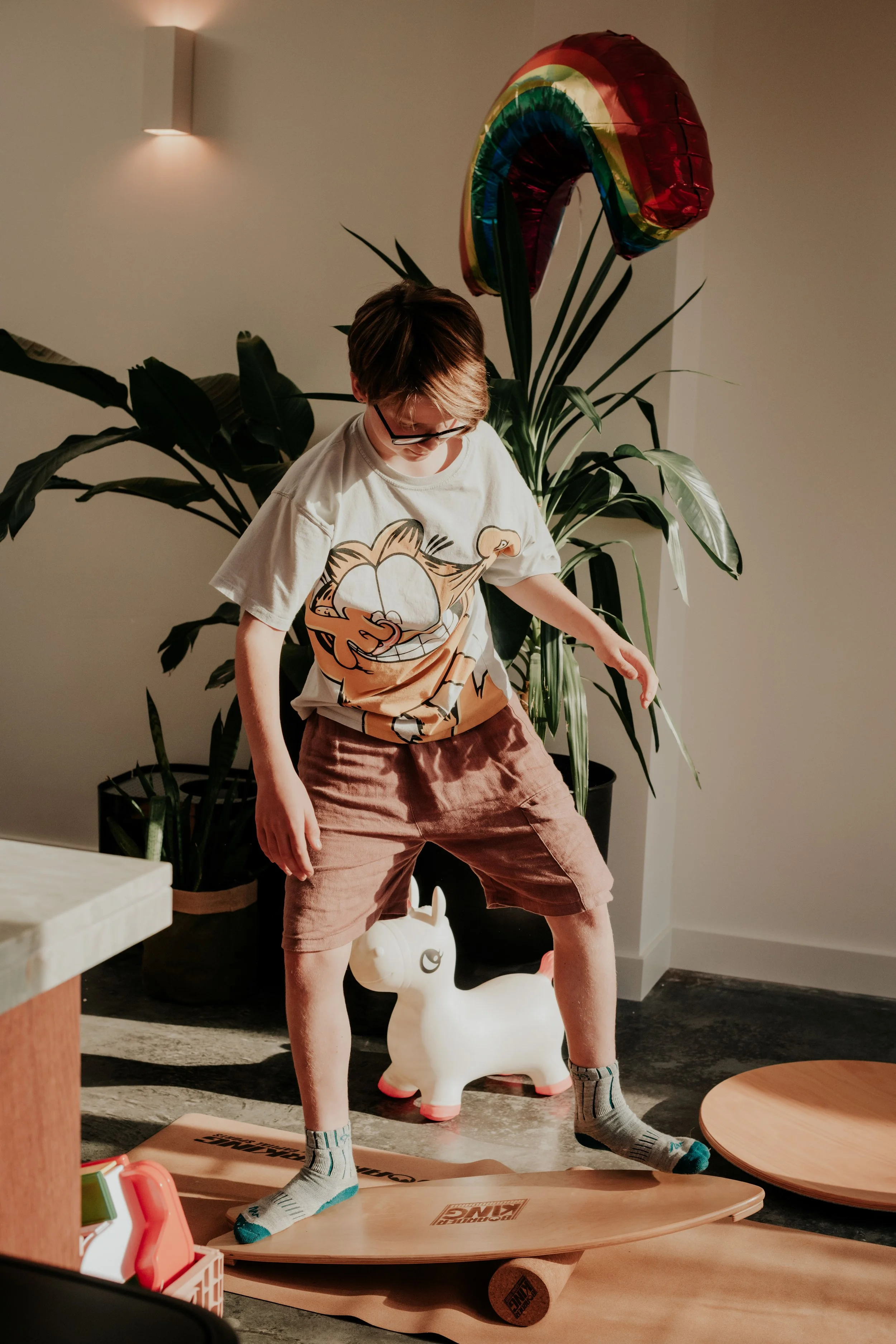A boy standing on a skateboard indoors near a large potted plant, with a rainbow-shaped balloon in the background. He is wearing glasses, a graphic t-shirt, brown shorts, and socks.
