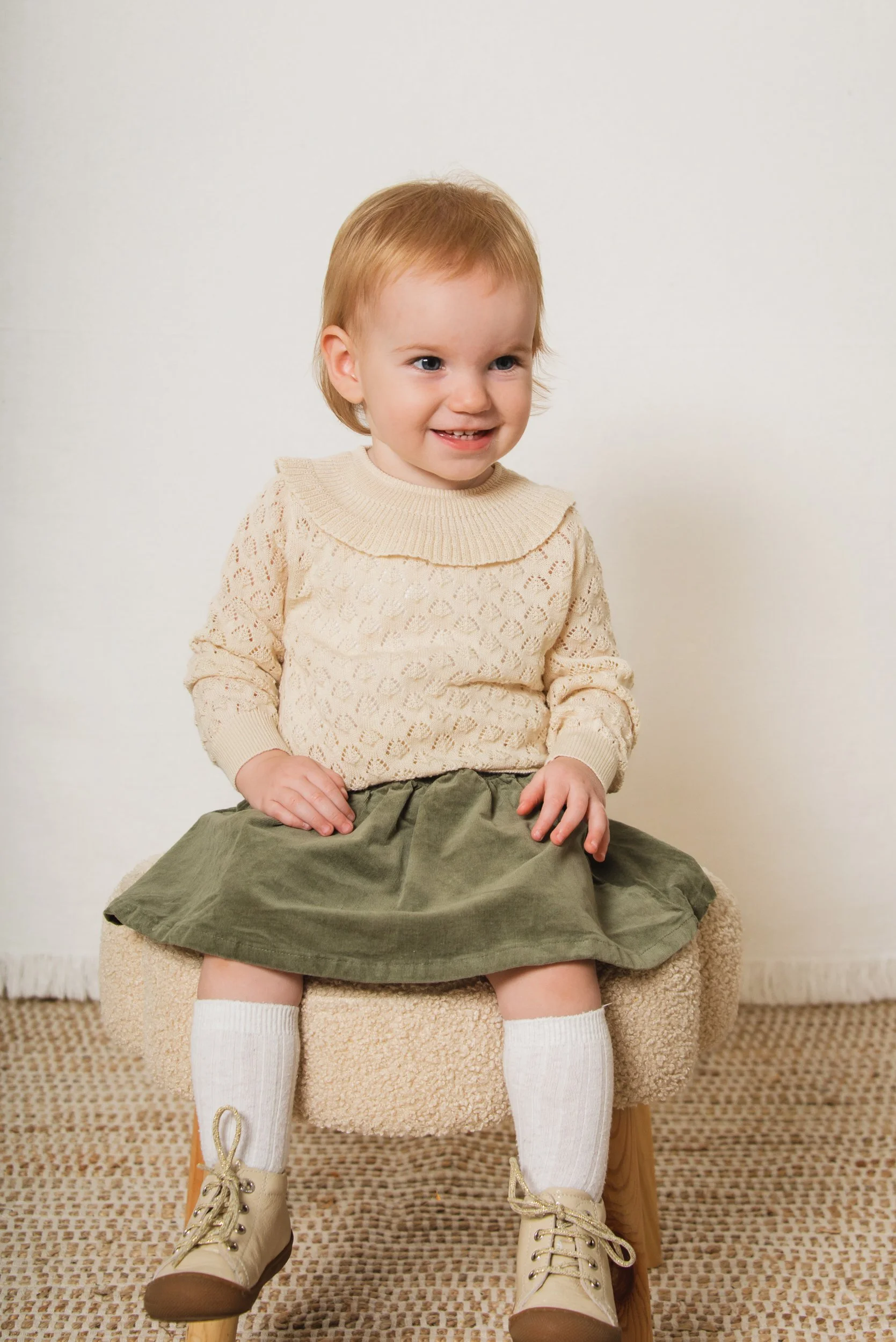 A young girl with red hair smiling while sitting on a beige chair in a room with a neutral background. She is wearing a cream lace sweater, a green skirt, white knee-high socks, and beige boots with laces.