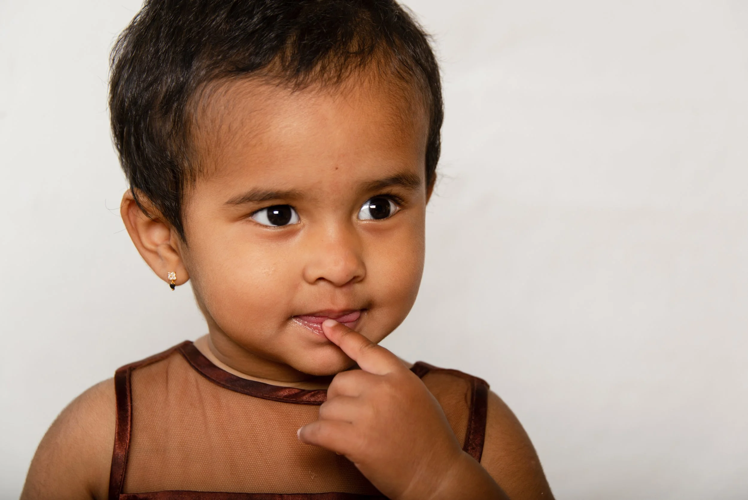 A young girl with dark hair, brown eyes, and earrings, wearing a sleeveless brown top, resting her finger on her lips and looking contemplative against a plain white background.