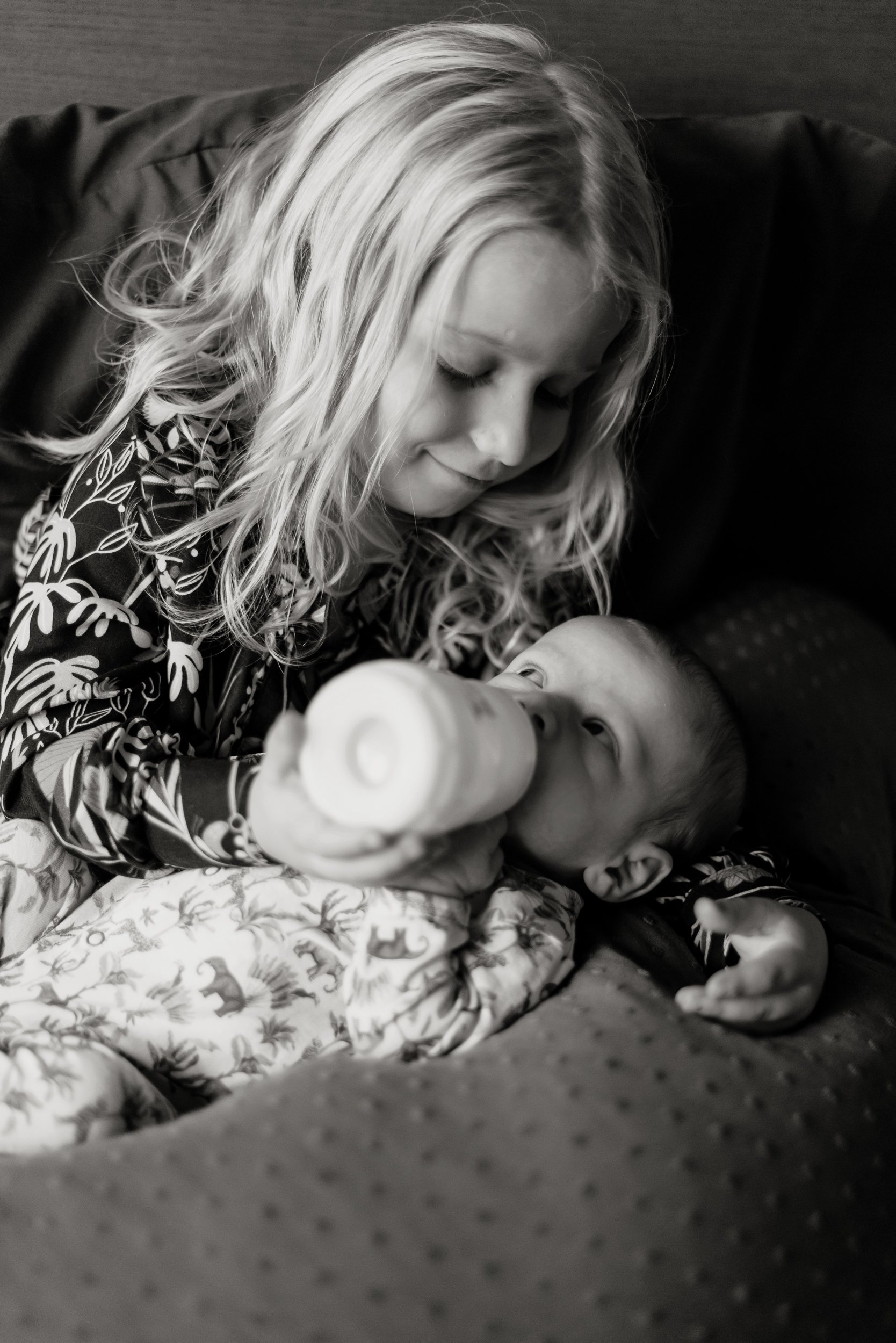 A woman with long curly hair feeding a young child with a bottle. The woman has her eyes closed and is looking down at the child, who is lying on a patterned blanket, wearing pajamas with animals on them.