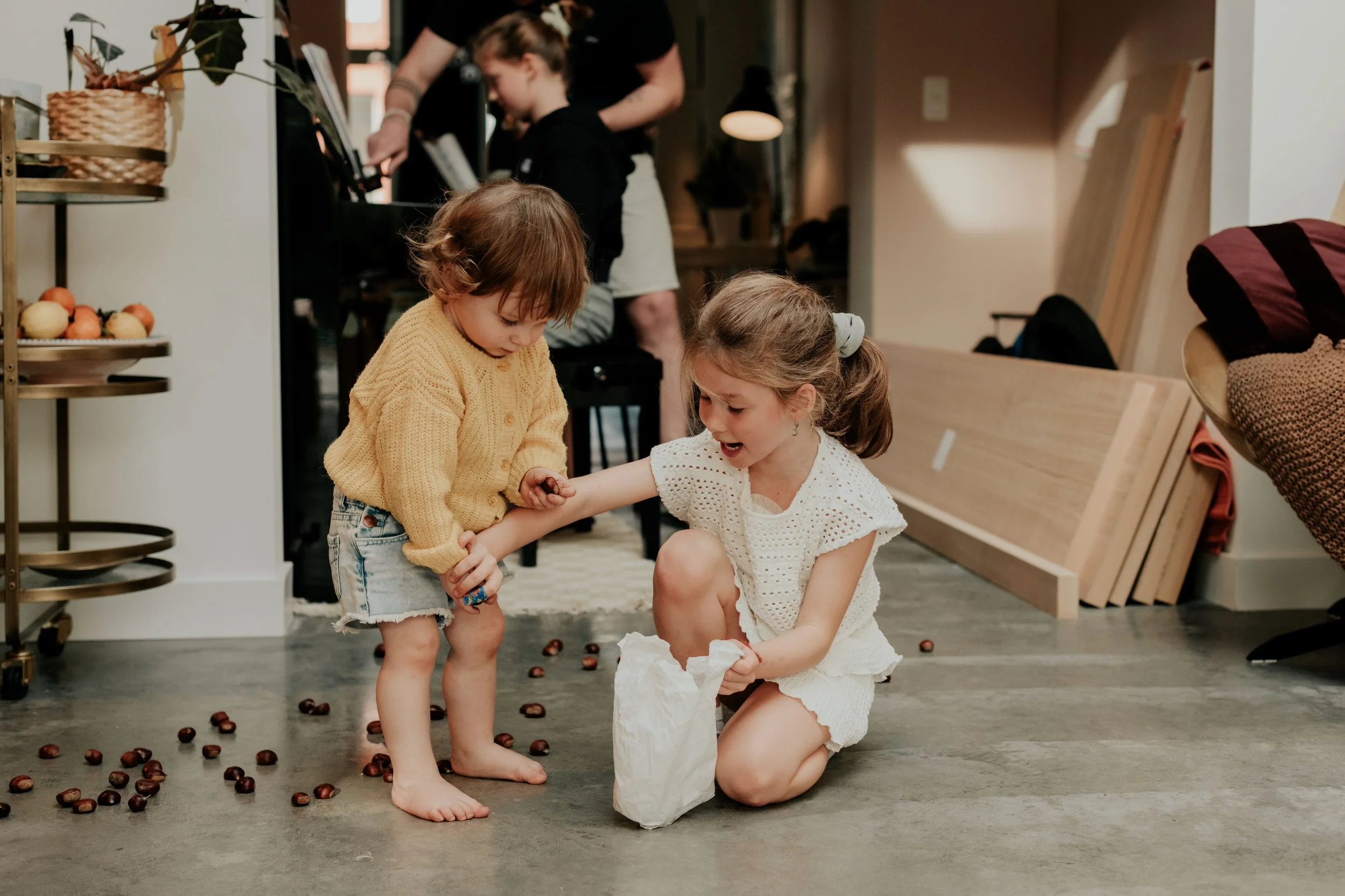 Two young girls are on the floor in a cozy living room, playing with chestnuts on the ground. One girl, wearing a yellow sweater and denim shorts, is standing while the other girl, dressed in white, is kneeling and holding a paper bag. There are peop