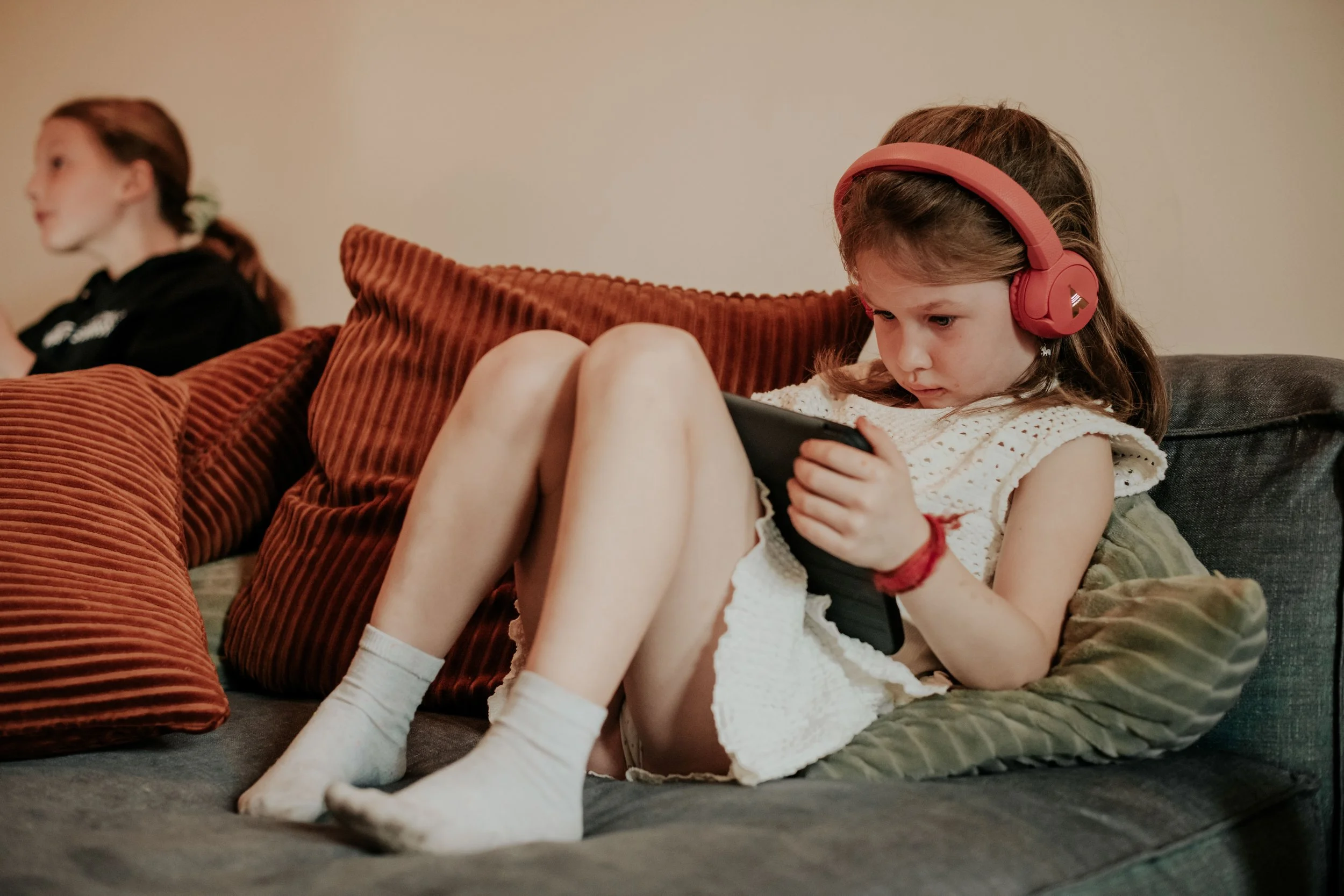 A young girl with brown hair wearing pink headphones is sitting on a couch, looking at her phone. She is dressed in a white dress with lace and striped knee-high socks. A woman with red hair tied back and wearing a black top is sitting next to her, f
