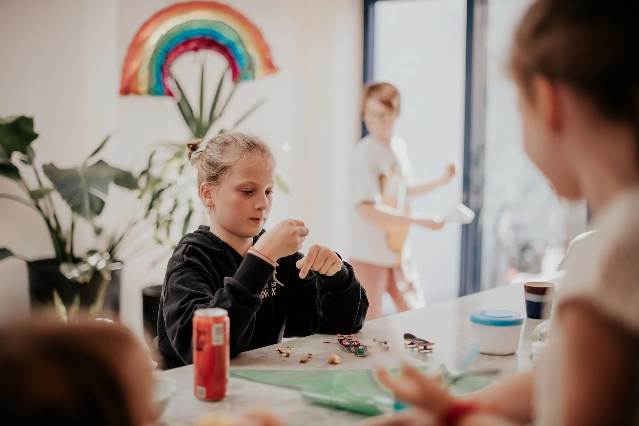 A group of kids sitting at a table arts and crafts activities, with a girl in a black hoodie in focus, working on a project. In the background, a woman is standing near a door, wearing glasses and a white shirt, smiling.