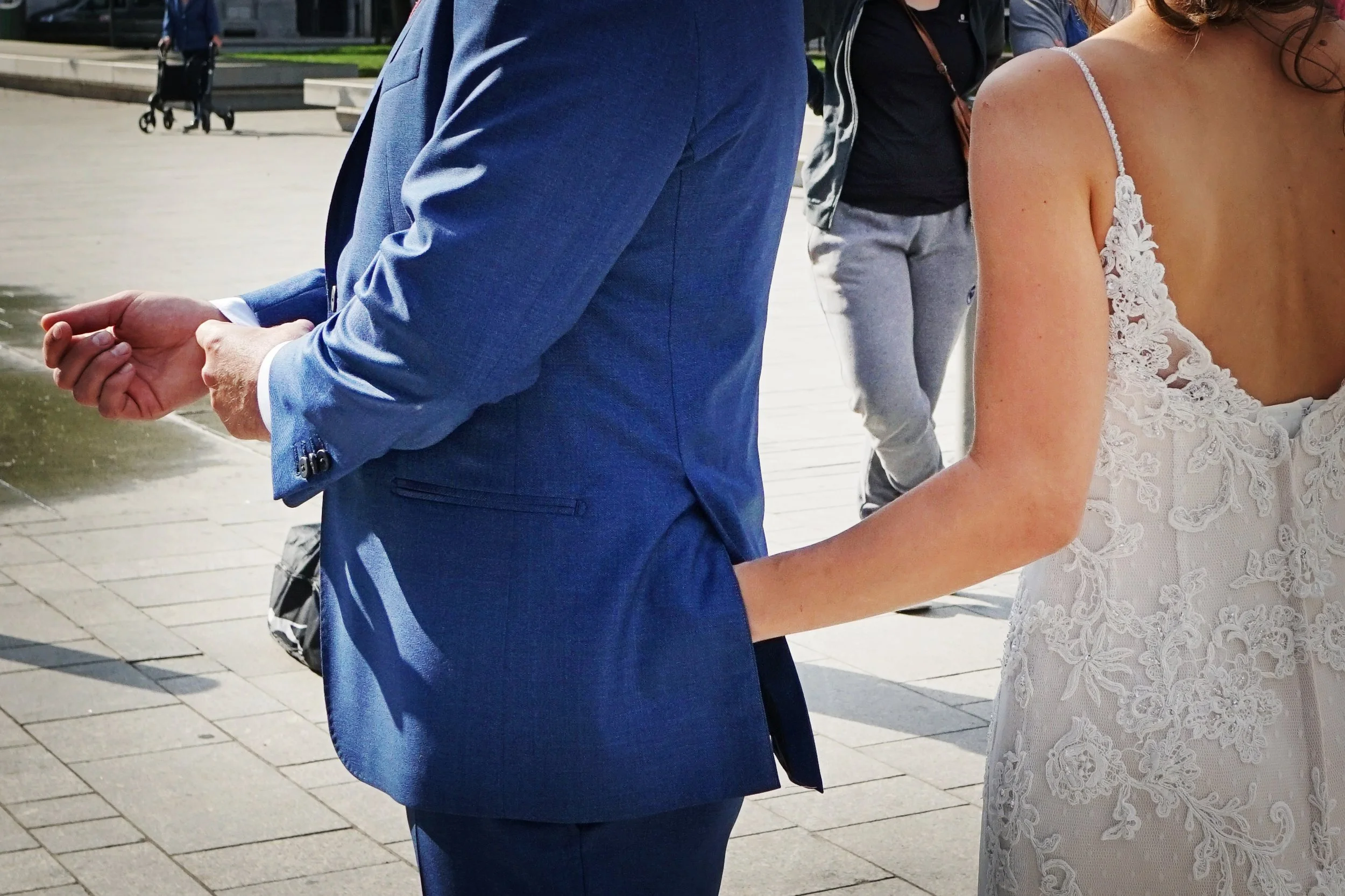 Close-up of a man in a blue suit and a woman in a white lace dress holding hands, with a blurred background of a woman walking with a stroller.