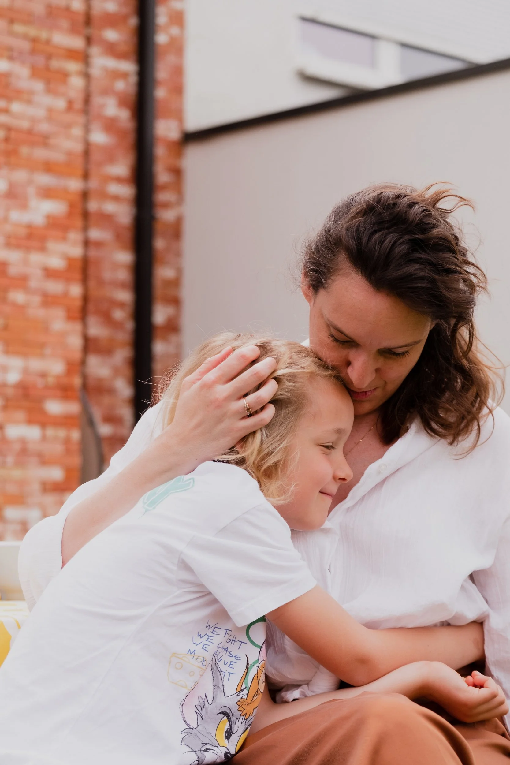 A woman with curly brown hair embracing and comforting a young girl with blonde hair, both wearing white shirts, in an outdoor setting with a brick wall and modern building in the background.