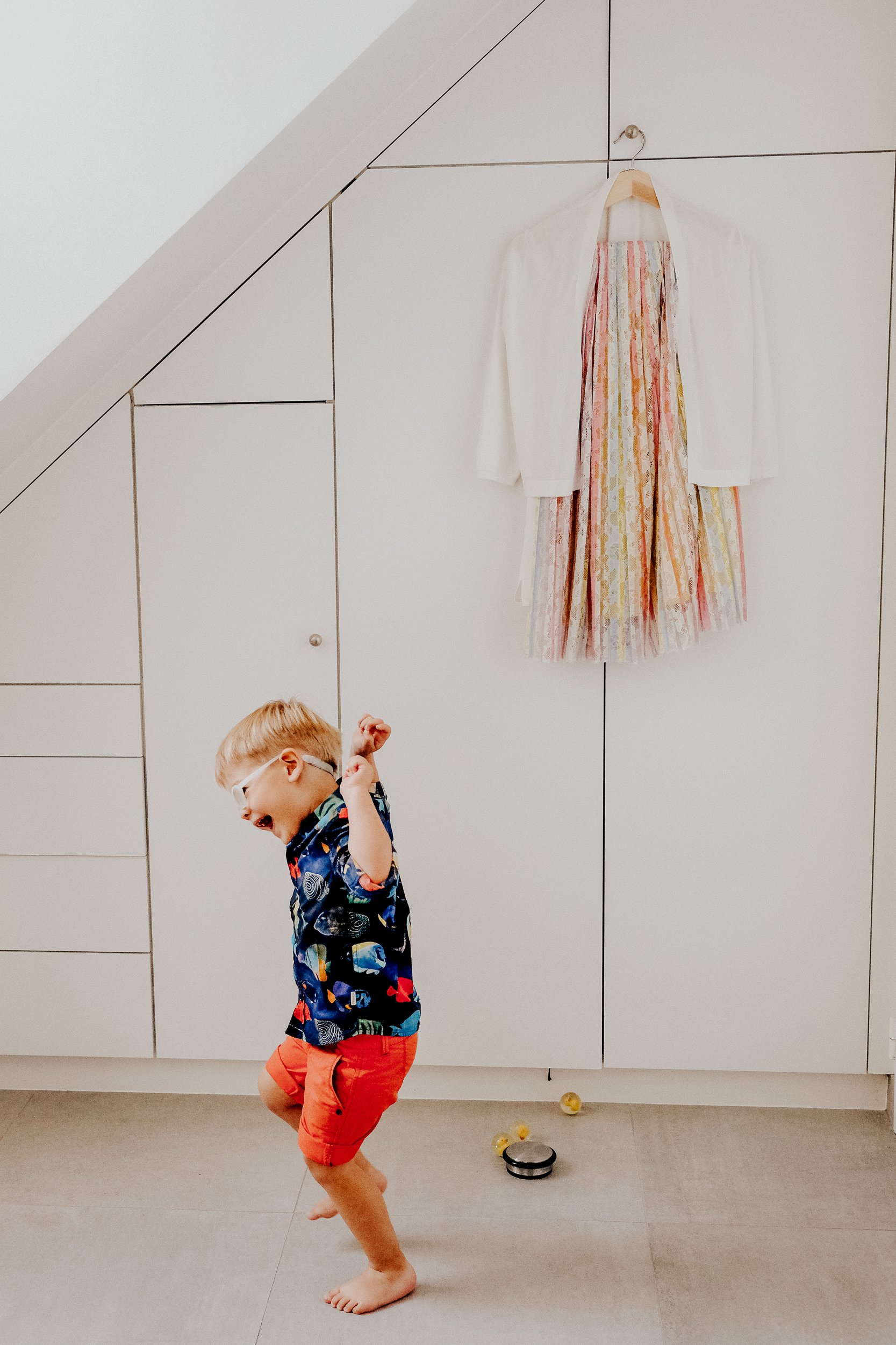 A young boy with glasses and a patterned shirt dancing barefoot in a room with a white tiled wall and cabinet, with a dress hanging on the wall and a robotic vacuum on the floor.