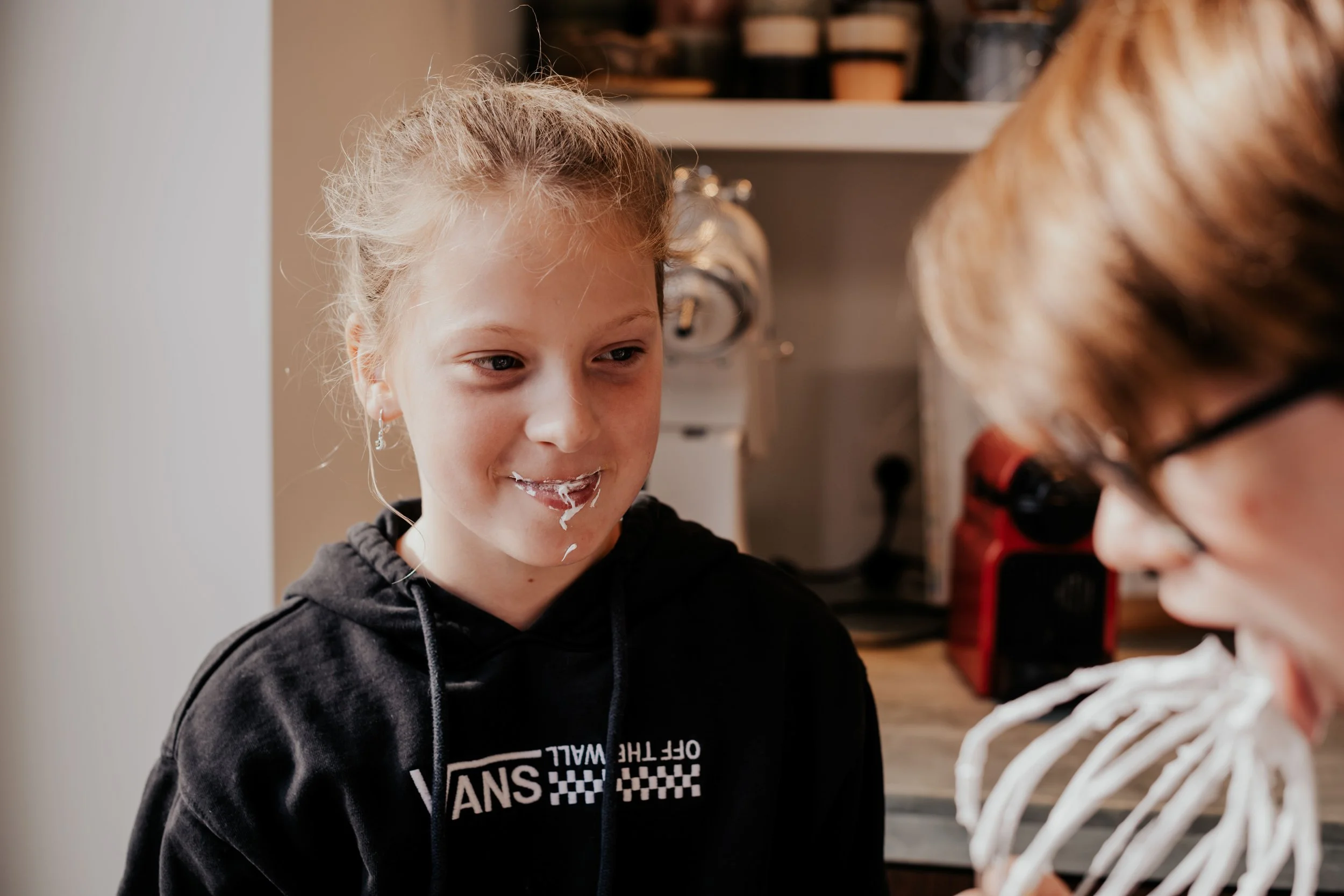 A young girl with blonde hair and earrings looks at a skeleton model with white teeth, in a room with shelves and equipment.