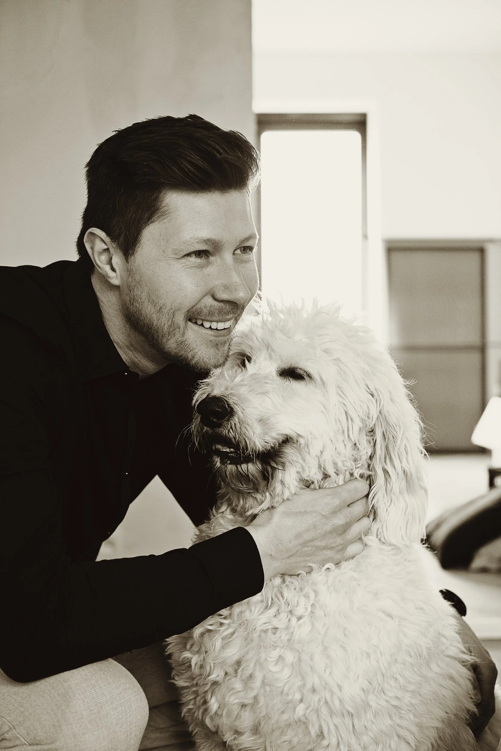 A man with dark hair and a beard smiling and hugging a large, curly-haired dog in a bright room.
