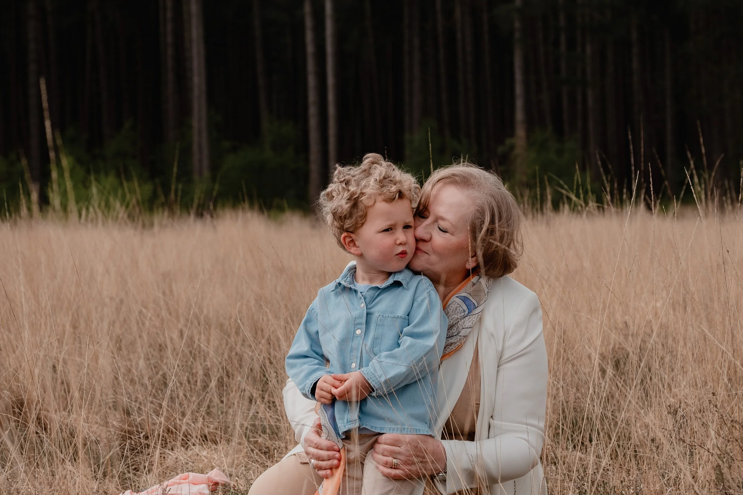 An older woman with short light hair kissing a young boy with curly blonde hair in a field of tall, dry grass, with a dense forest of dark trees in the background.
