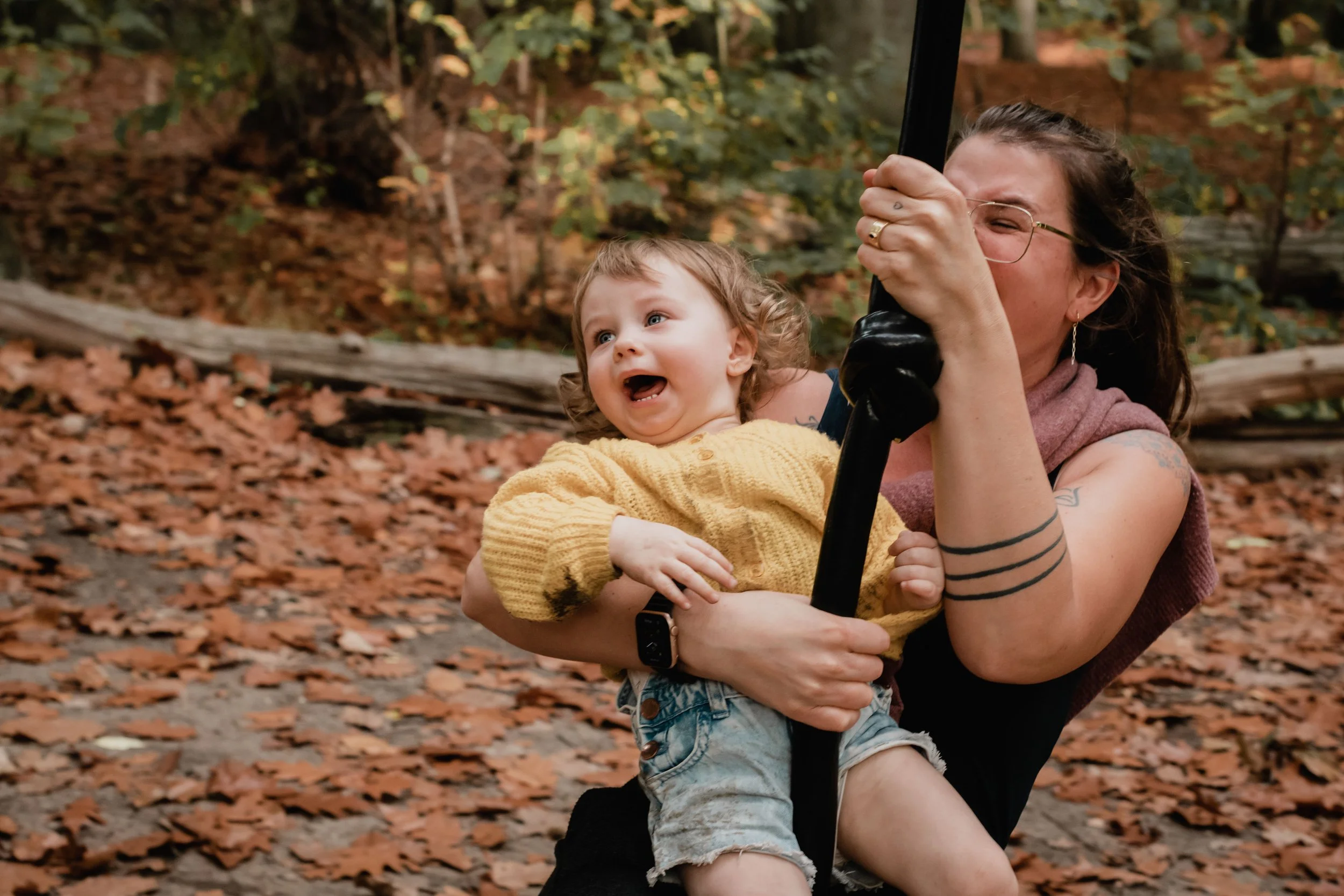 A woman with glasses and tattoos holding a young girl with curly hair and a yellow sweater in a forest with fallen leaves.