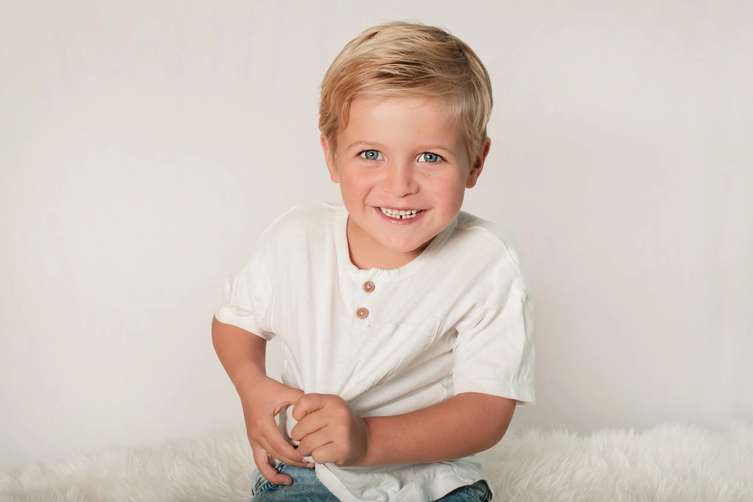 A young boy with blonde hair and blue eyes smiling, wearing a white shirt with buttons and blue jeans, sitting on a white furry surface against a plain light background.