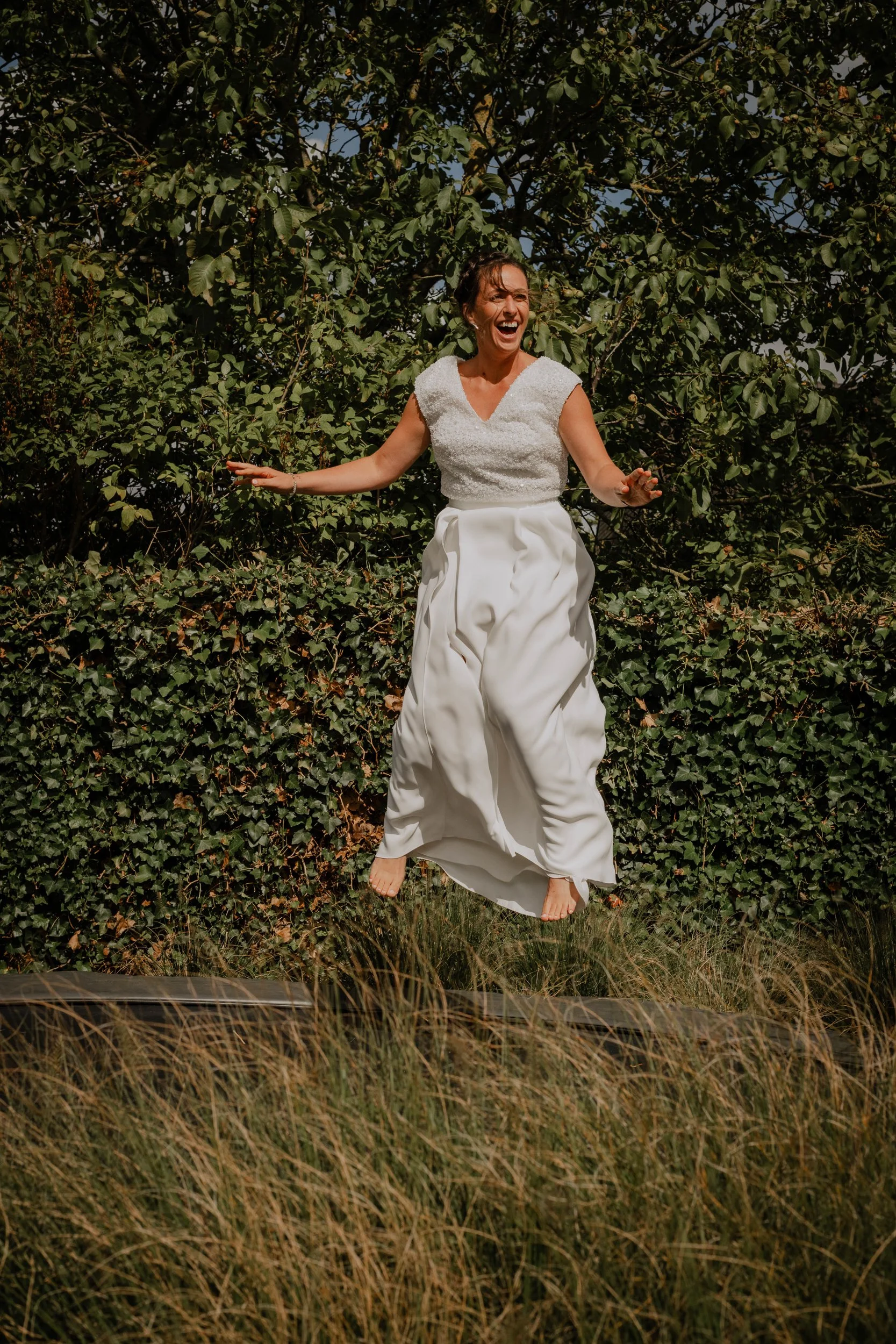 A woman in a white dress is jumping in the air outdoors, with green trees in the background and tall grass at her feet.