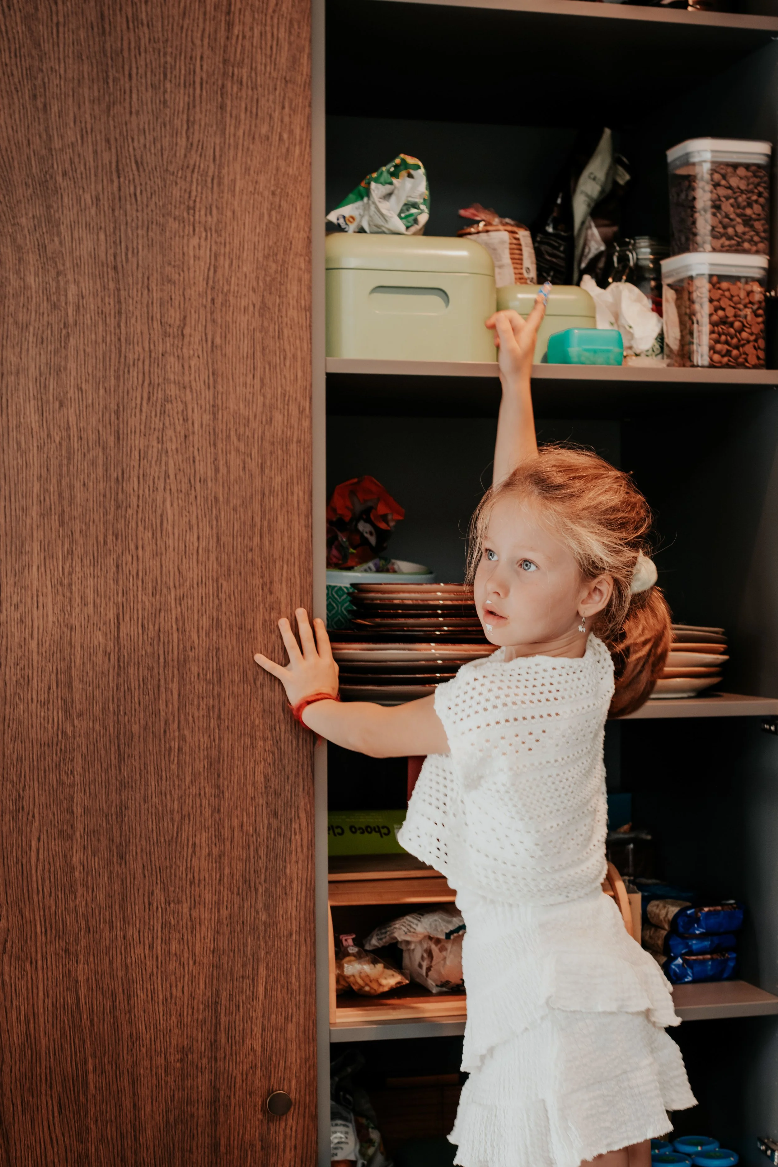 A young girl with curly red hair wearing a white dress is reaching into a dark-colored kitchen cabinet with her left hand. The cabinet is open, revealing food items and containers inside. The girl is looking upward and has a curious expression.