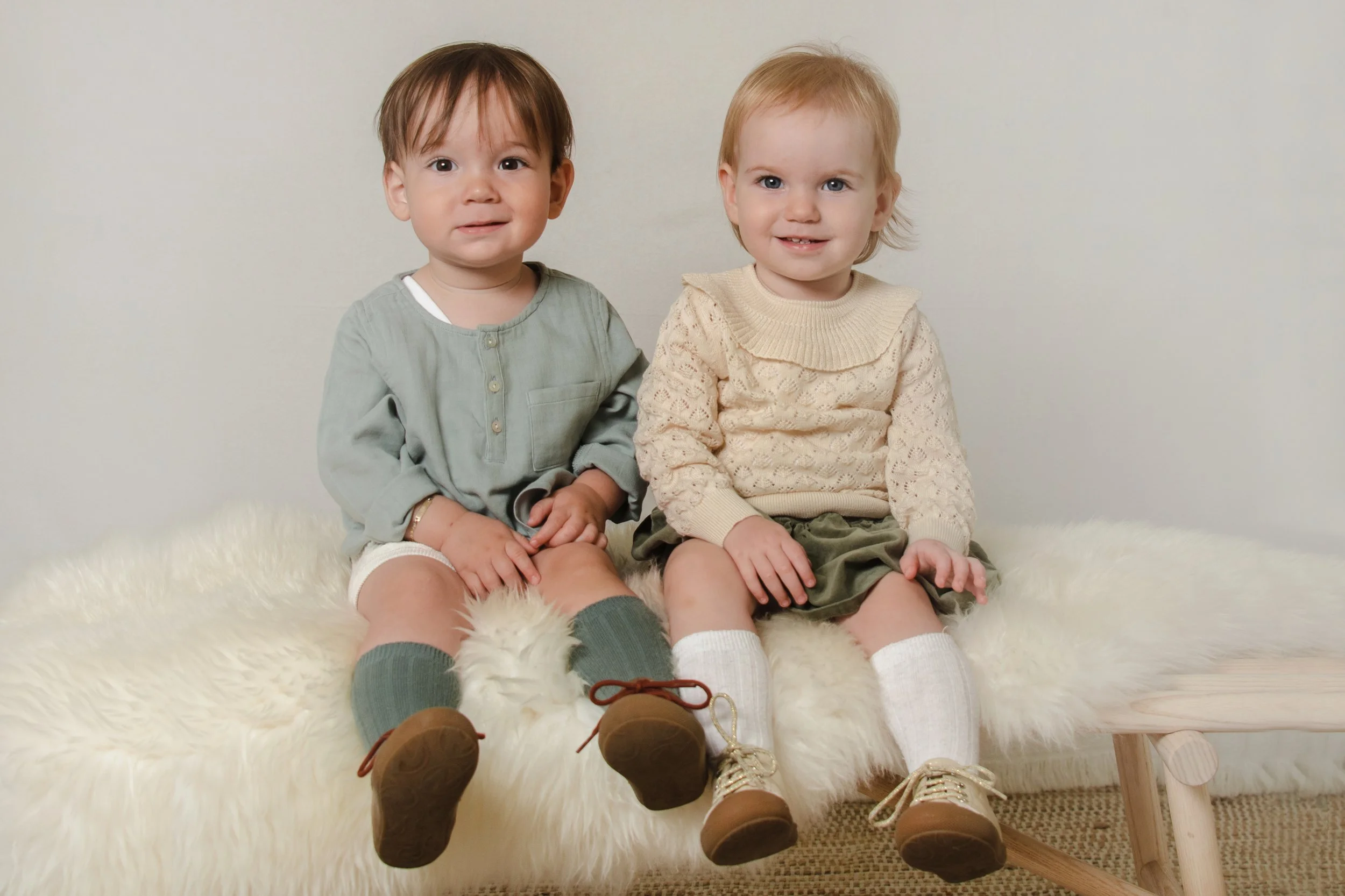 Two young children sitting together on a fluffy white rug, facing the camera, against a plain light-colored wall. The child on the left has brown hair and a light green shirt, while the child on the right has blonde hair and a cream-colored sweater. 