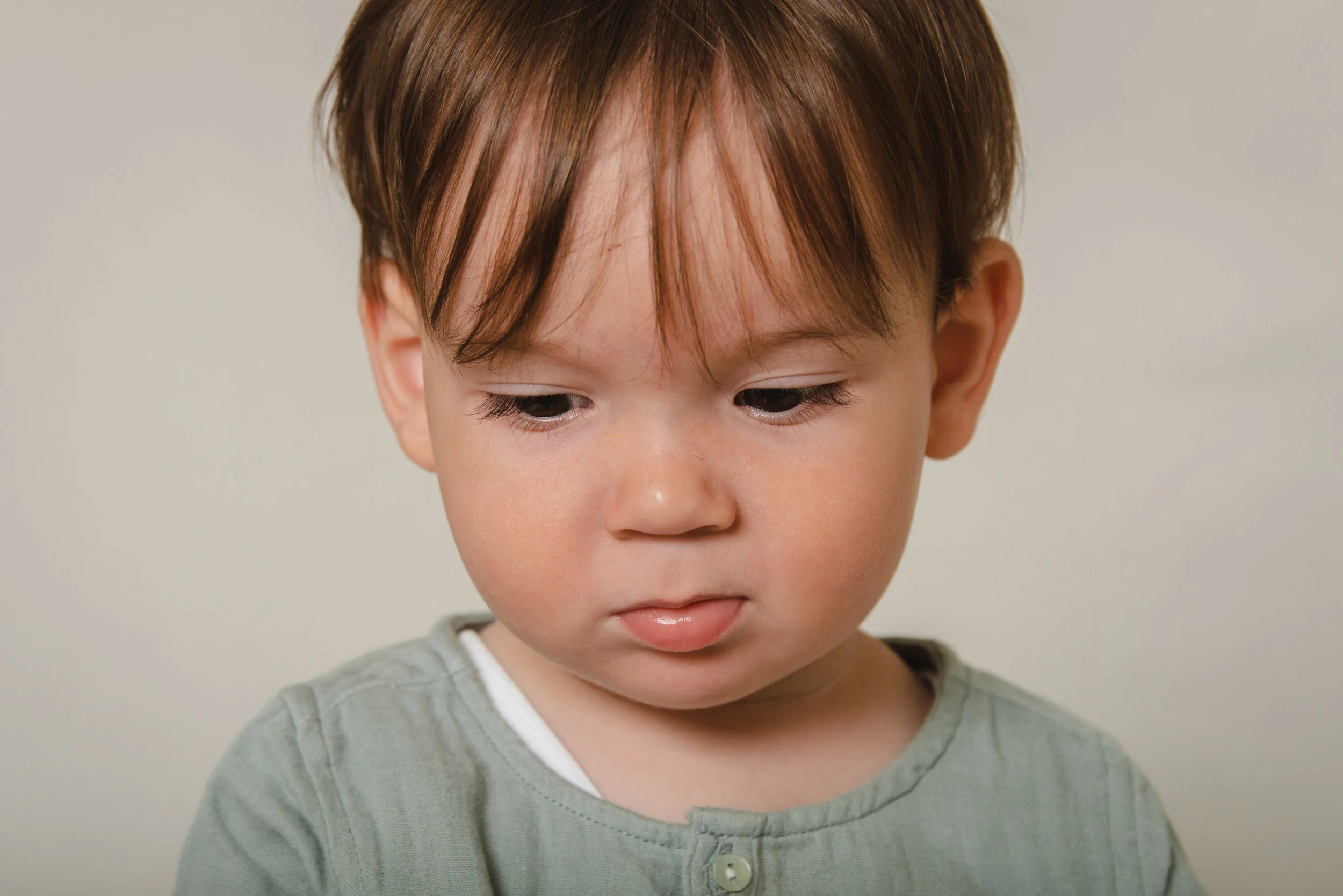 Close-up of a young boy with brown hair, wearing a green shirt, looking down against a plain light gray background.
