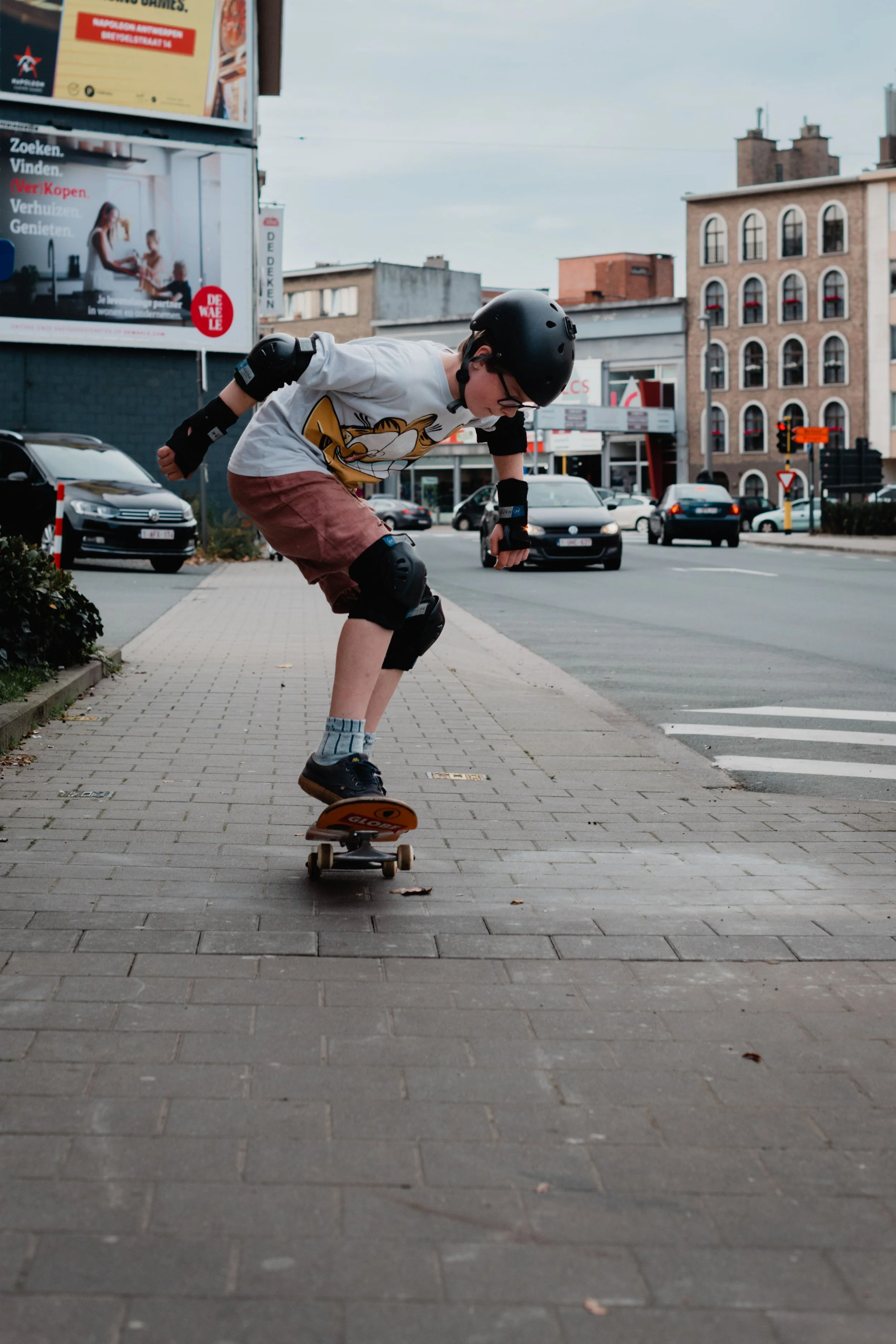 A young person wearing a helmet, glasses, and protective gear is riding a skateboard on a city sidewalk. They are leaning forward, focusing on their skateboarding.