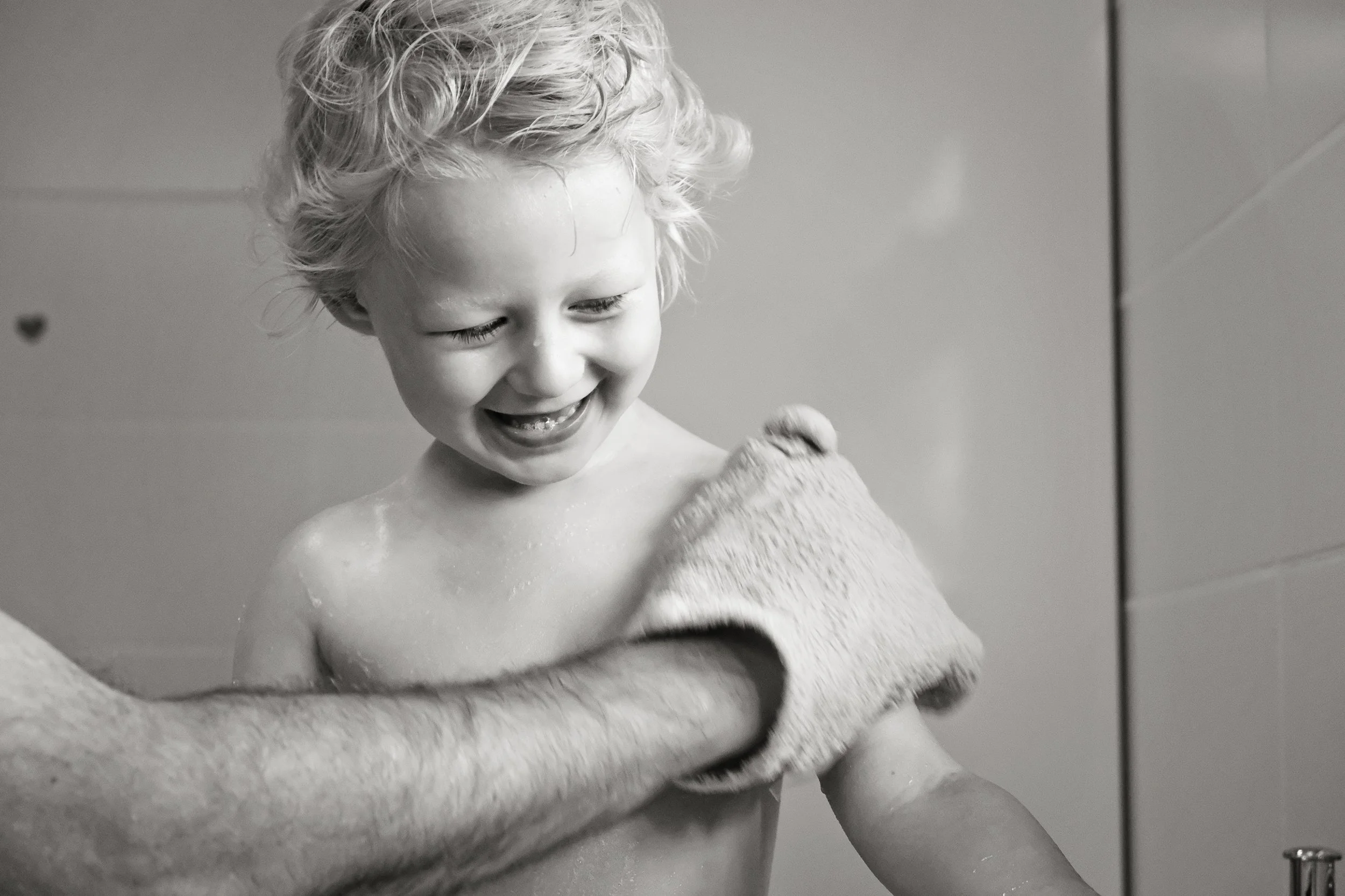 A young child with curly hair smiling happily during a bath, wrapped in a towel.