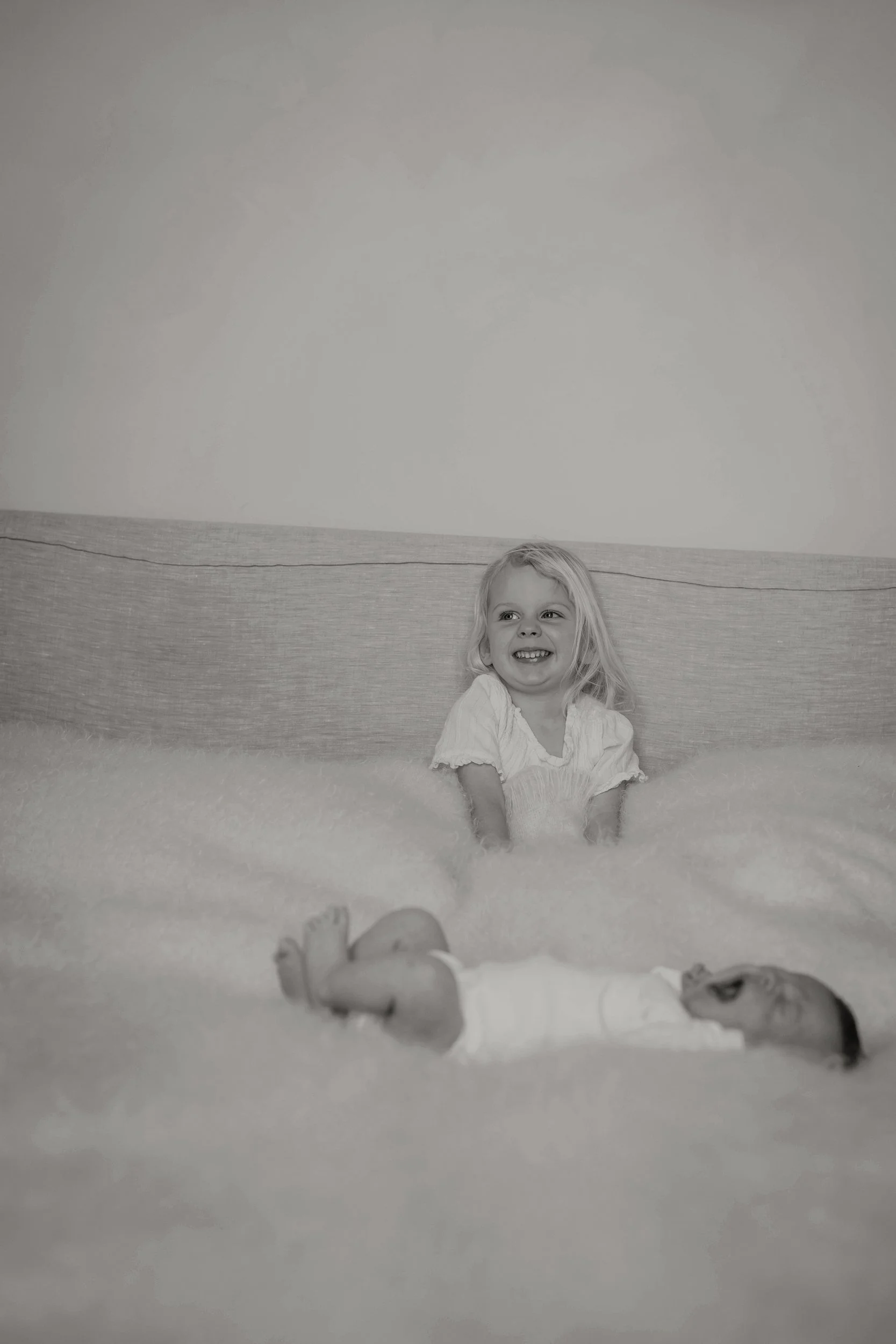 Black and white photo of a smiling young girl sitting on a blanket with her legs stretched out in front of her. The girl is wearing a light-colored dress, and her hair is shoulder length. There is a plush dog toy lying beside her on the blanket. The 