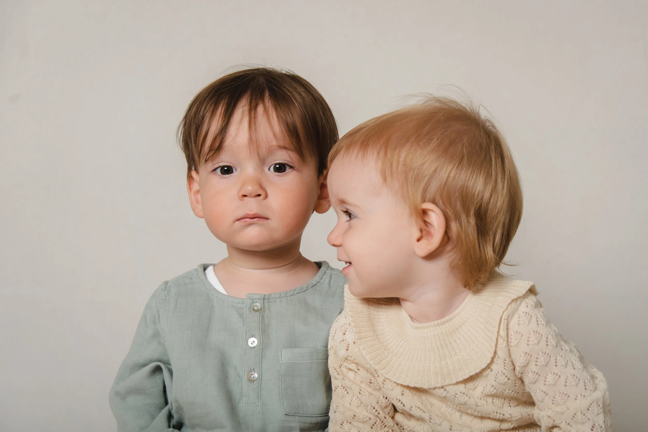 Two young children, a boy and a girl, standing close together against a plain, light-colored background. The boy has brown hair and a serious expression, while the girl has red hair and is turned towards the boy, smiling.