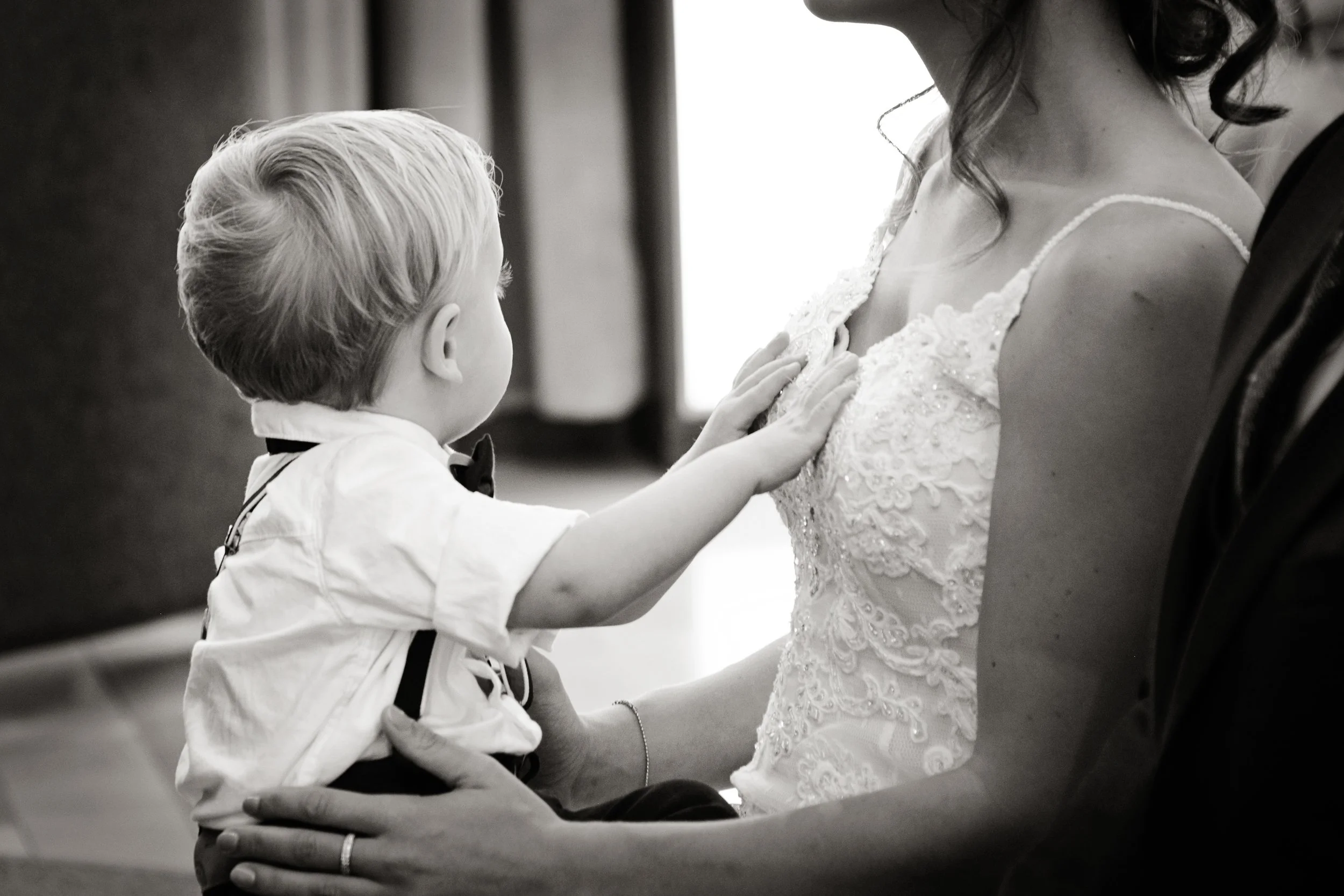 A woman wearing a wedding dress with lace details and a young boy in formal attire, including a bow tie, touching her wedding dress gently.