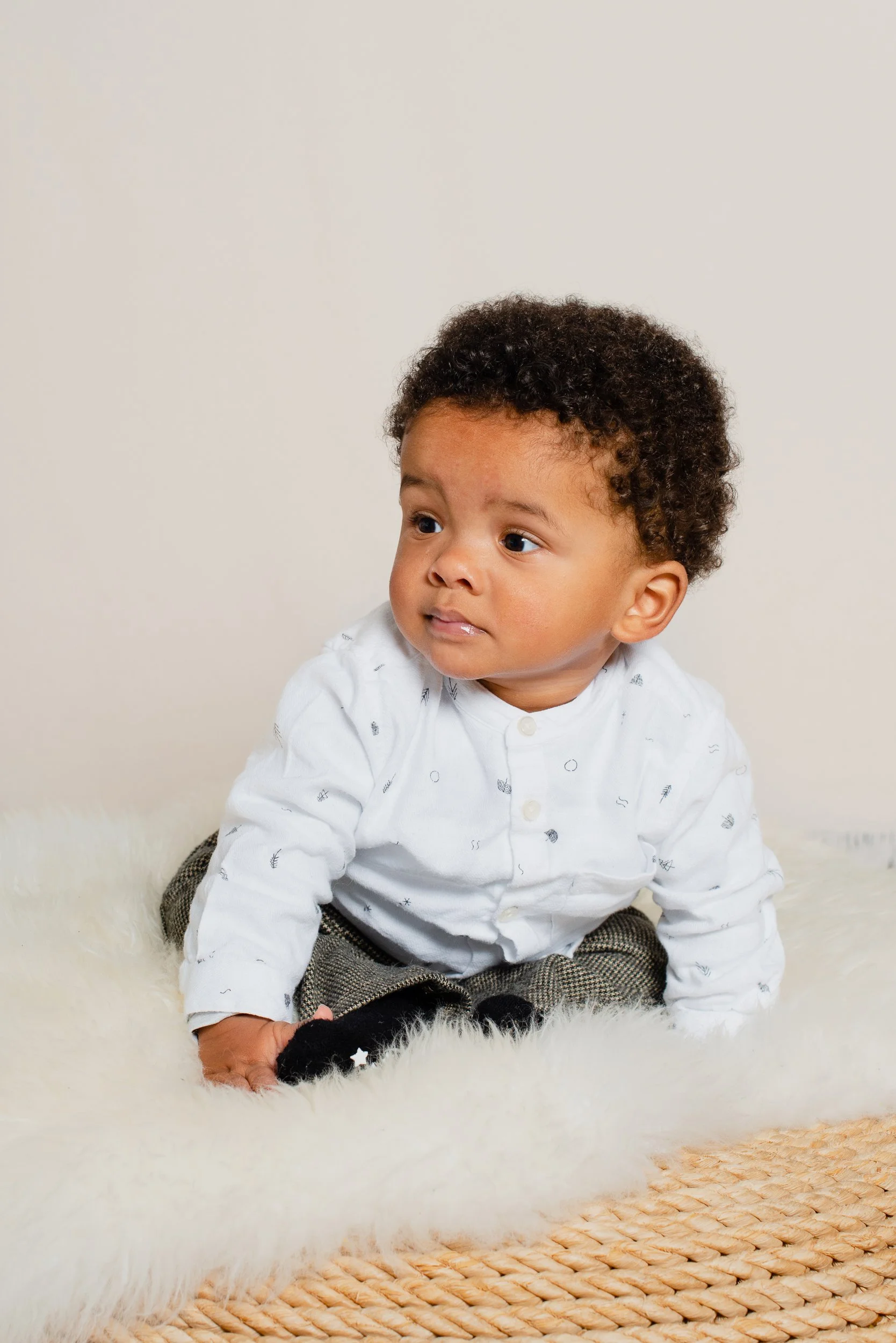 A young boy with curly hair, wearing a white shirt, sitting on a fluffy white rug, in front of a plain light-colored wall.