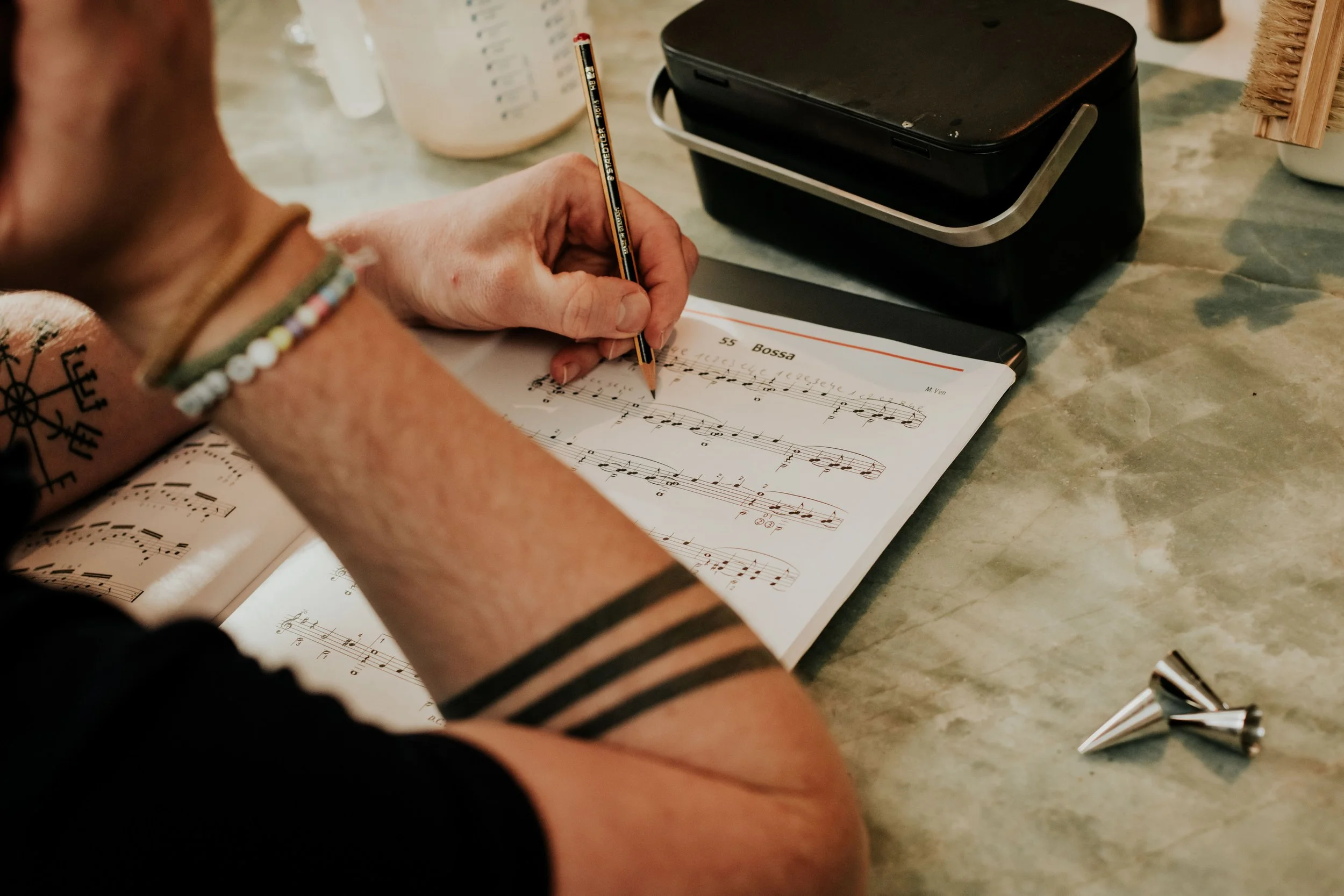 A person wearing a beaded bracelet and a black armband is writing musical notes on sheet music titled "Bossa" with a pencil. There is a printer, a pen, and decorative items on the marble table.