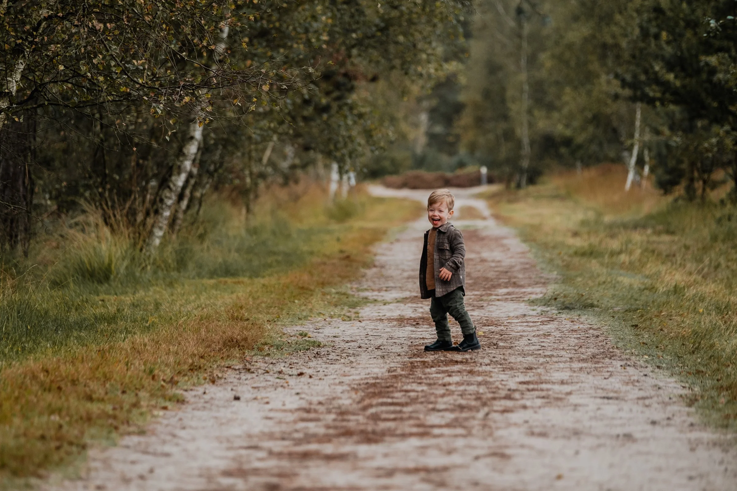 A young boy walking along a dirt path in a wooded area, smiling and dressed in a coat and boots during fall.