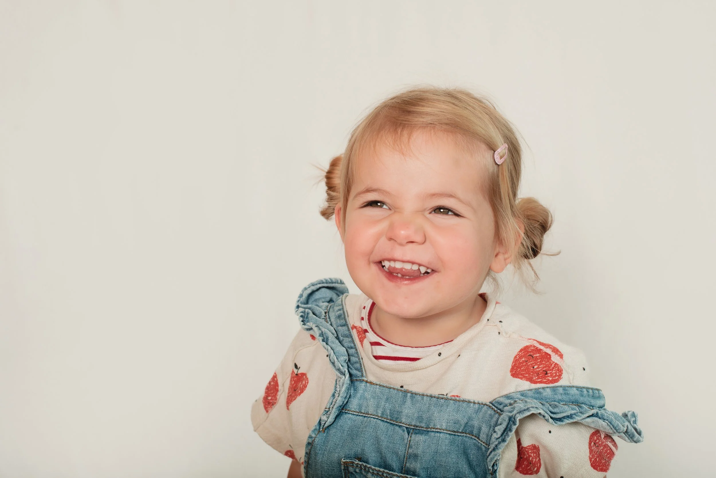 A young girl with blonde hair styled in pigtails, wearing a white shirt with red strawberries and denim overalls, smiling and looking slightly to her right against a plain light-colored background.