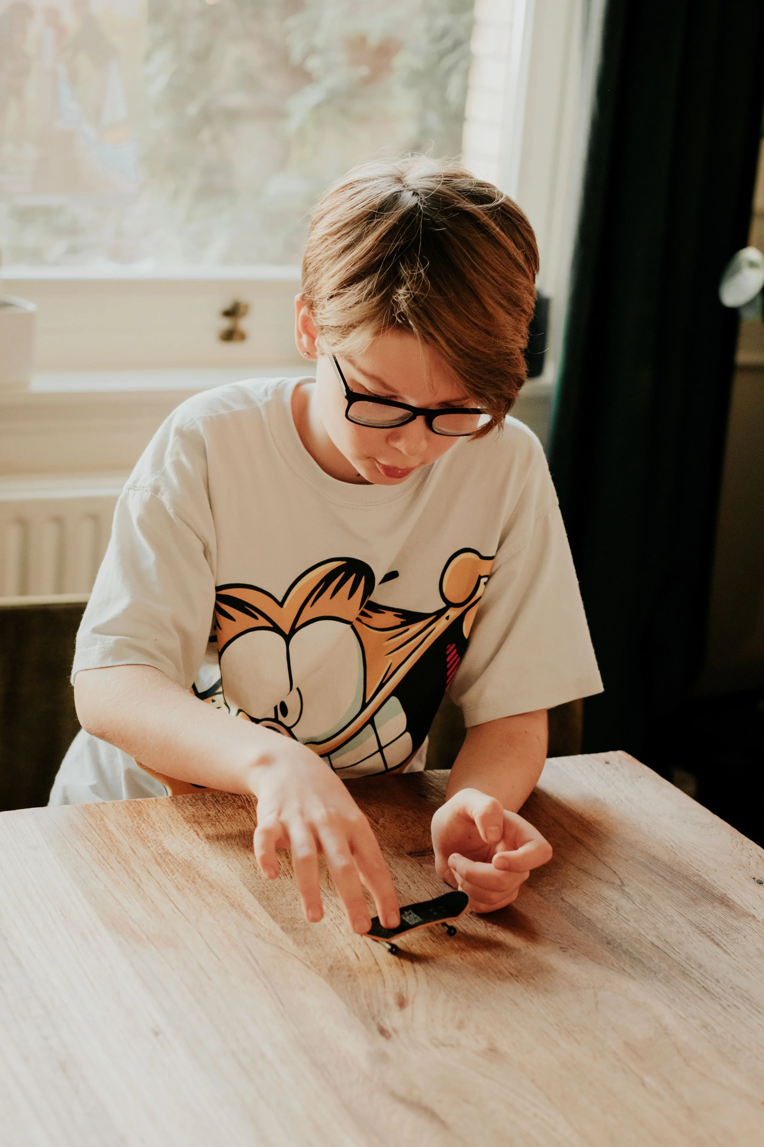 A person with glasses and short hair wearing a cartoon character t-shirt appears to be using a small tool or device on a light-colored wooden table beside a window with some greenery outside in the background.