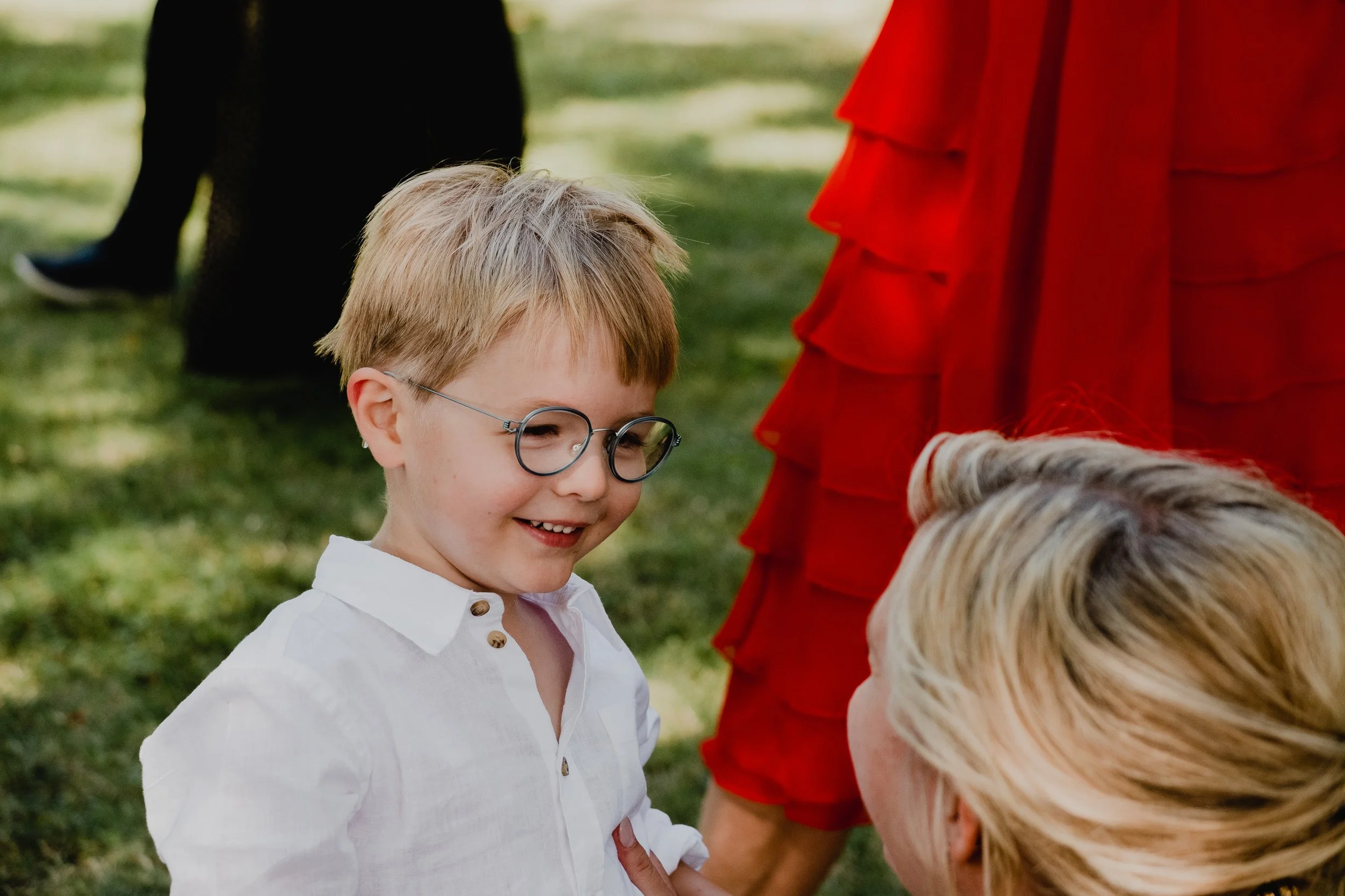 A young boy smiling and wearing glasses outdoors during the daytime, with other people around, including a woman in a red dress.