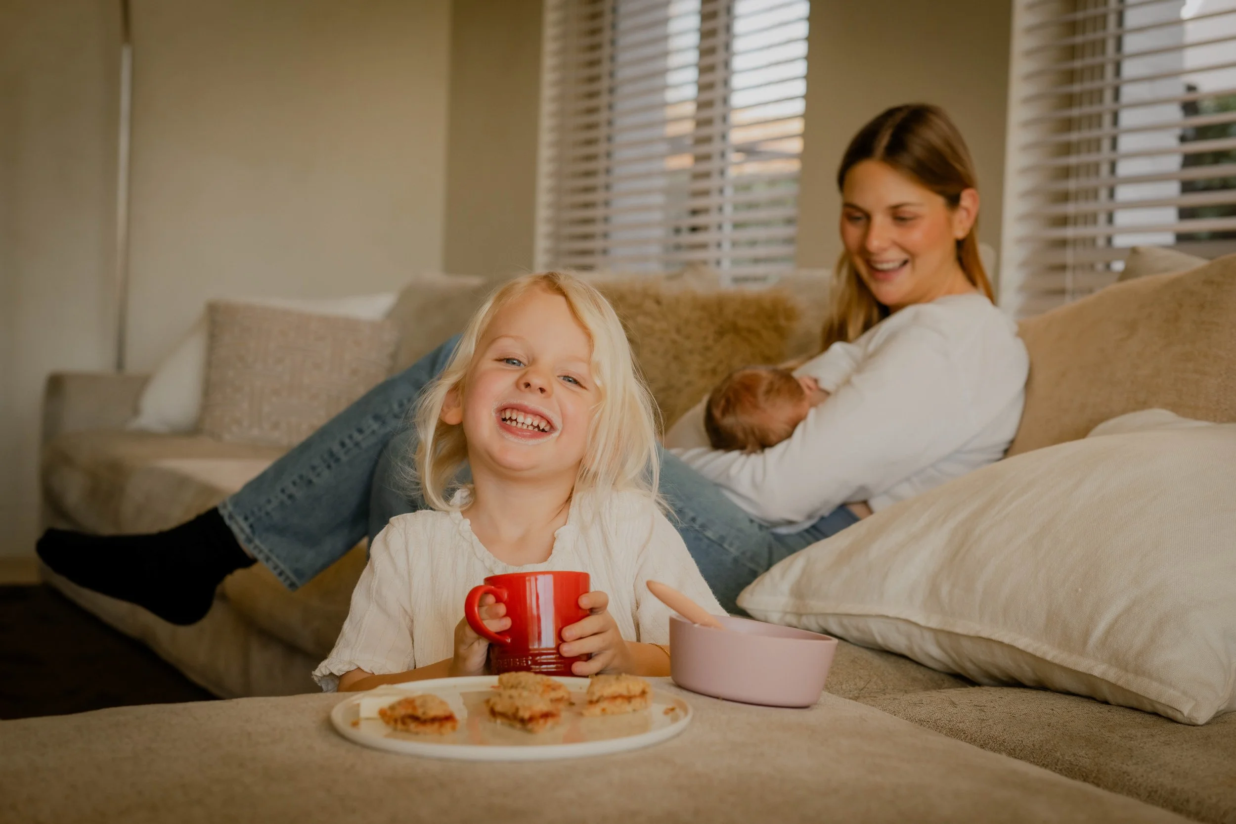 A young girl with blonde hair smiling and holding a red mug, sitting on the floor in front of a low table with pizza slices, with an adult woman and a newborn baby sitting on a sofa in the background.