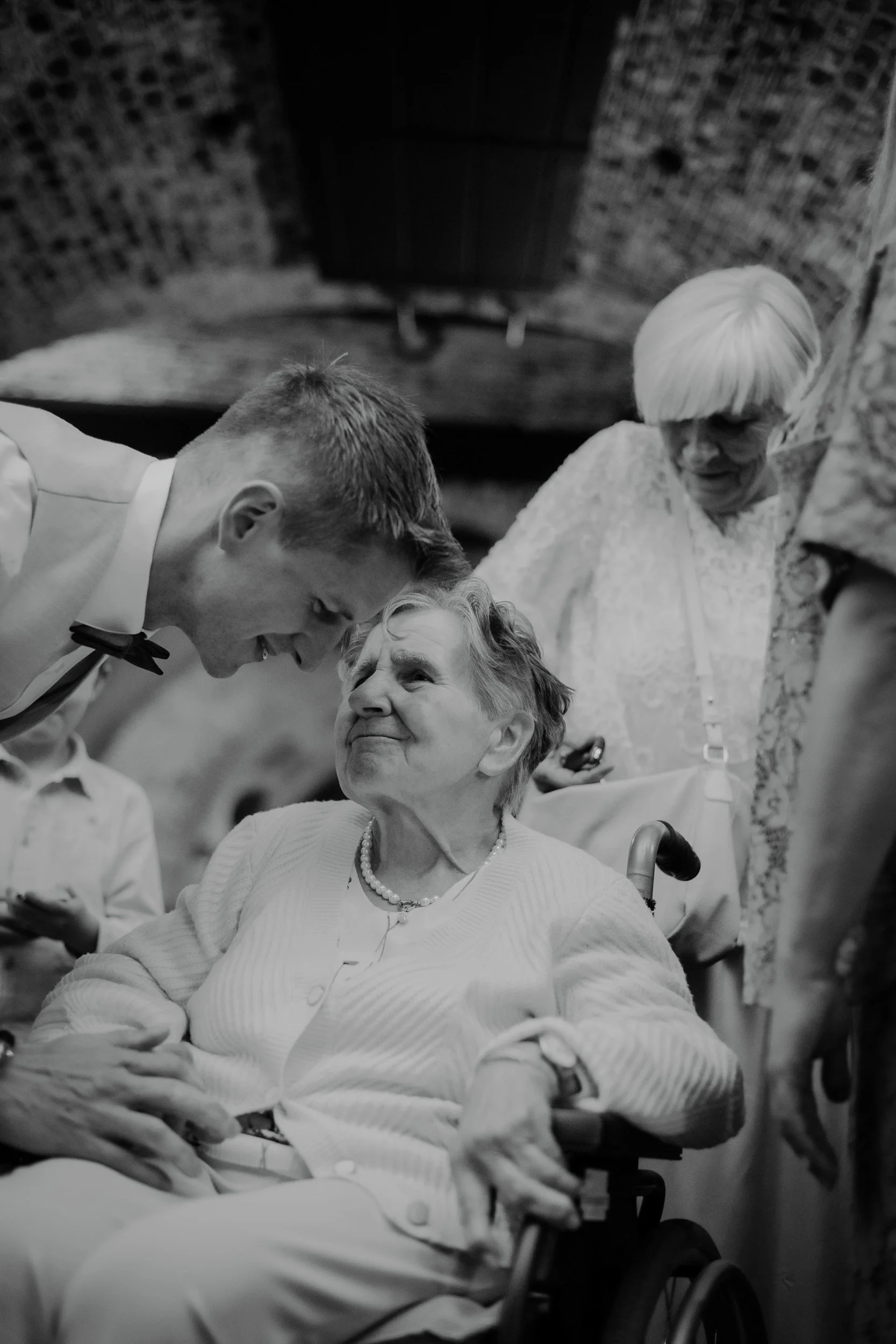 An elderly woman with short gray hair sitting in a wheelchair, smiling and looking up at a young man leaning toward her. Two women are standing behind her, one with white hair wearing a lace top and the other wearing a patterned dress. The scene appe