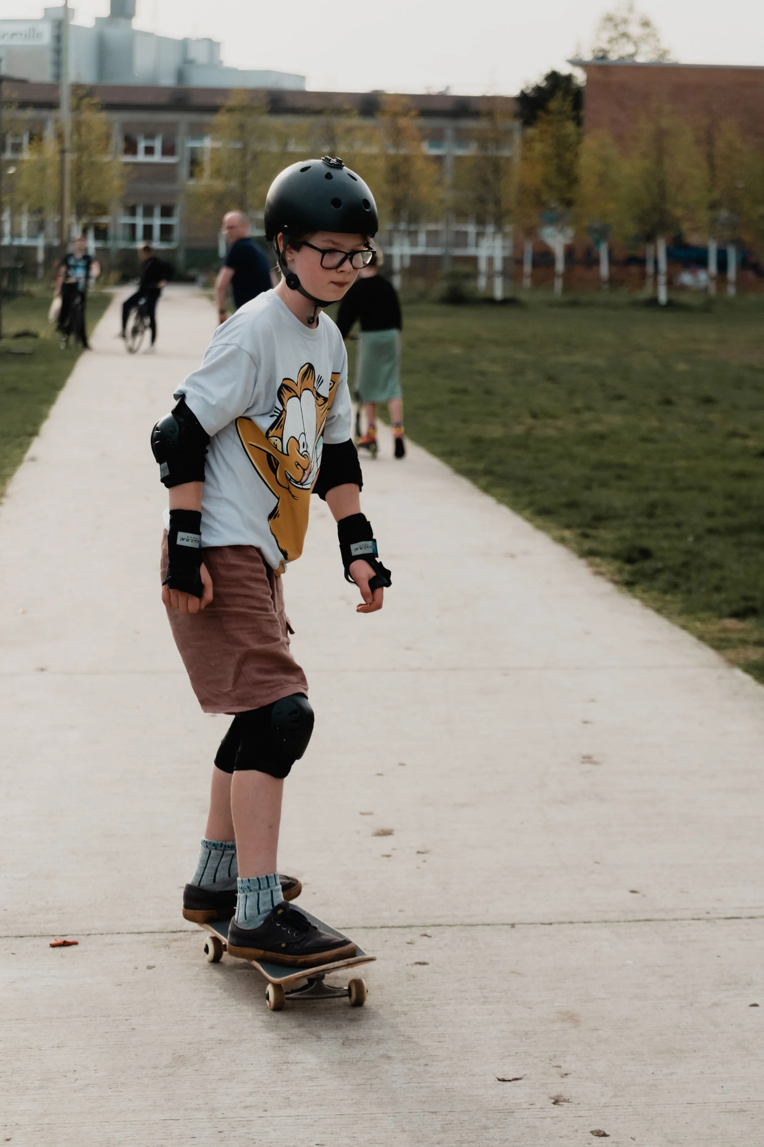 A young boy wearing glasses, a helmet, and elbow pads skateboarding on a paved path in a park, with several people and trees in the background.