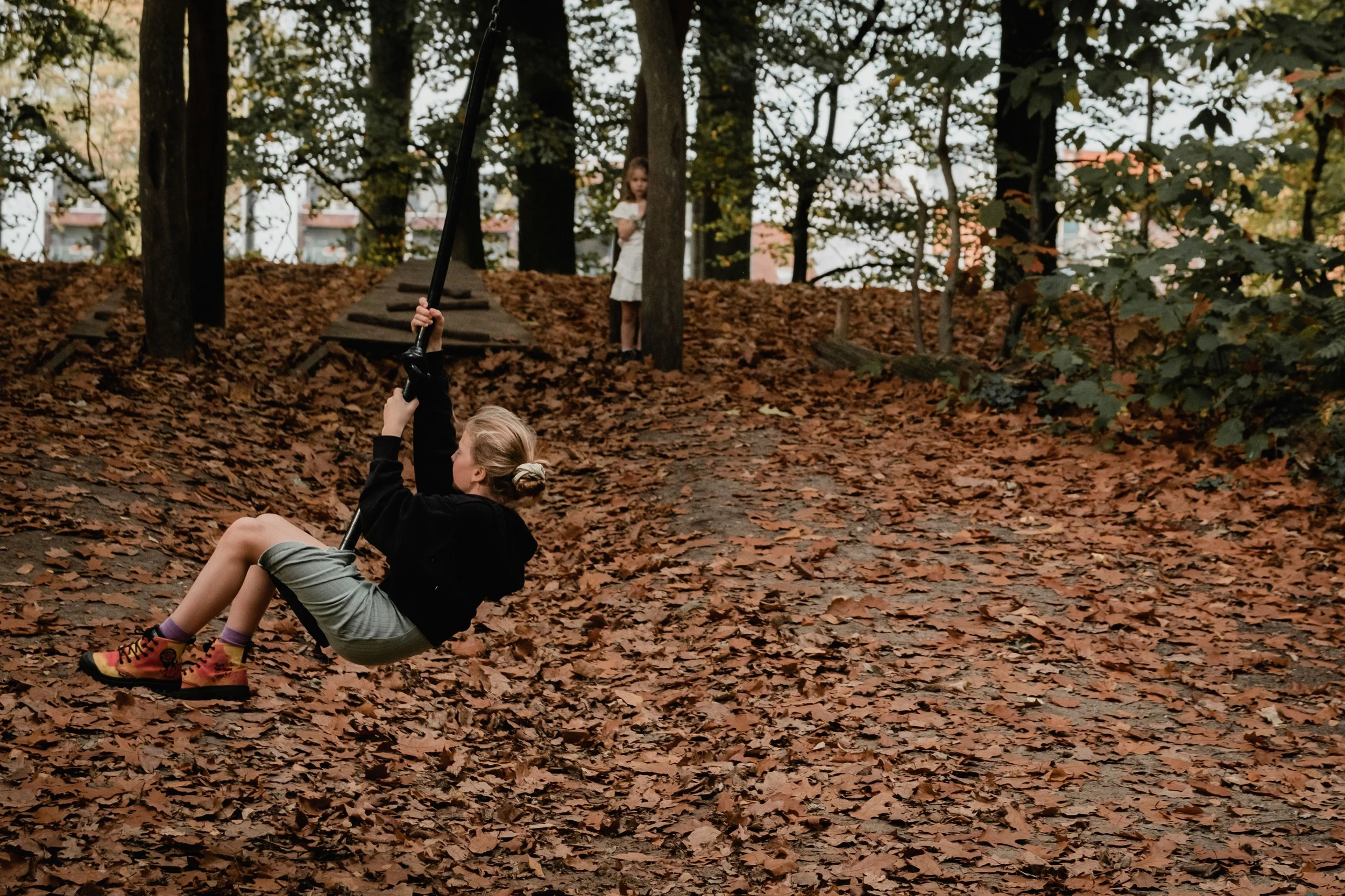 A girl sitting in fallen autumn leaves holding onto a swing with another girl behind a tree in a wooded park.