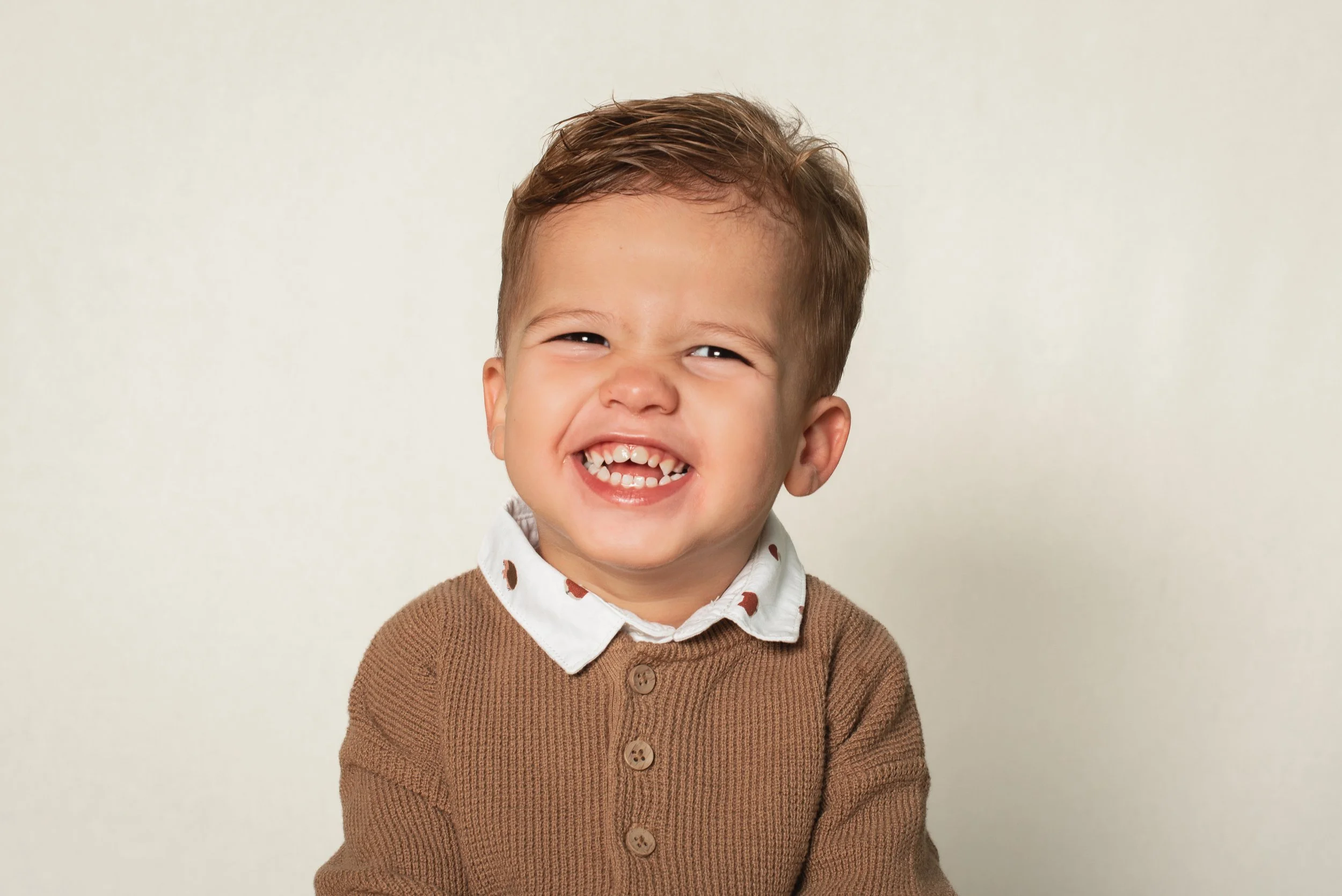 A young boy with brown hair smiling widely, showing his teeth, wearing a white collared shirt with a brown sweater, against a neutral background.