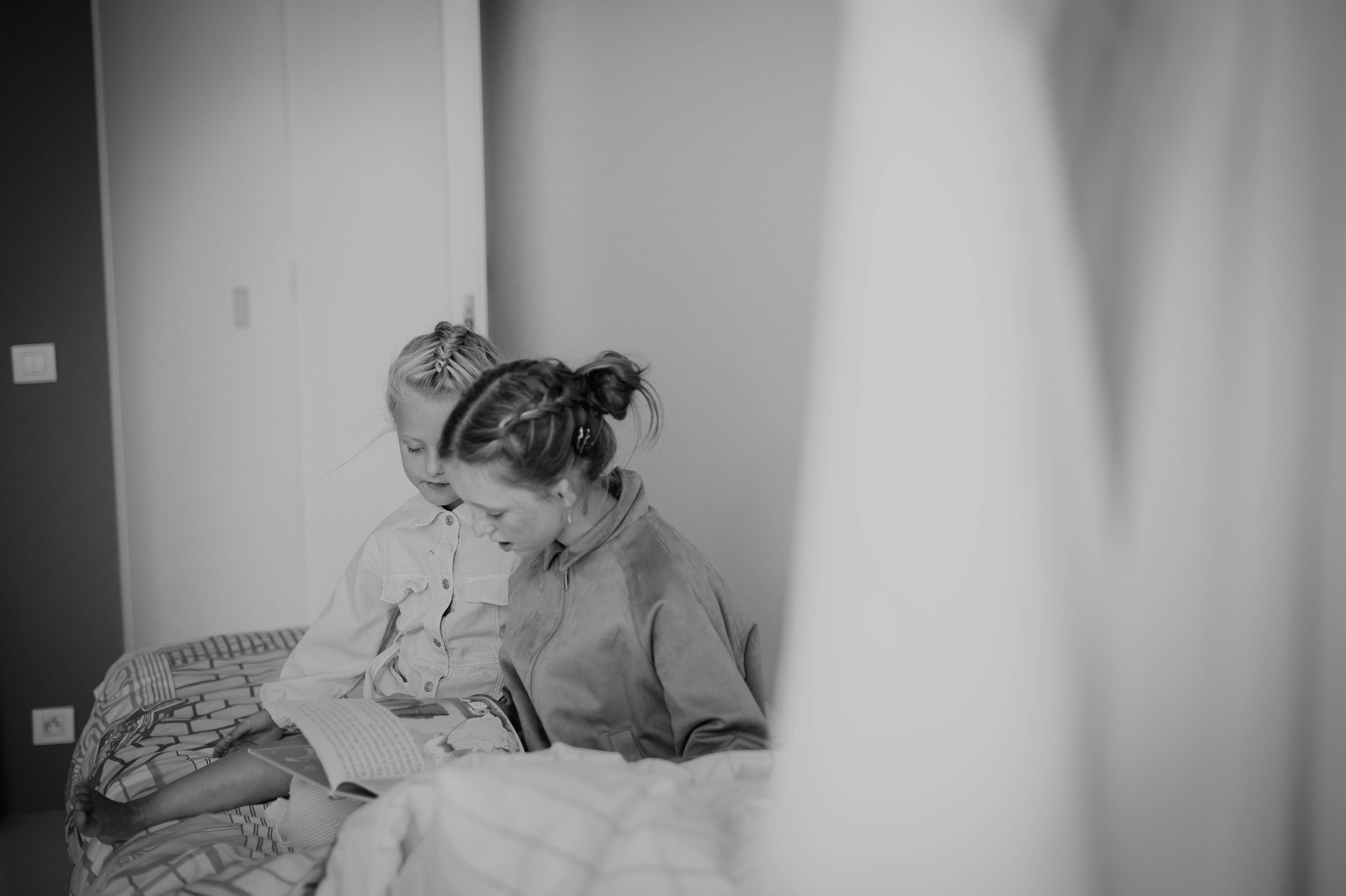 Two girls with braided hair sitting together on a bed, reading a book in a room with plain walls.