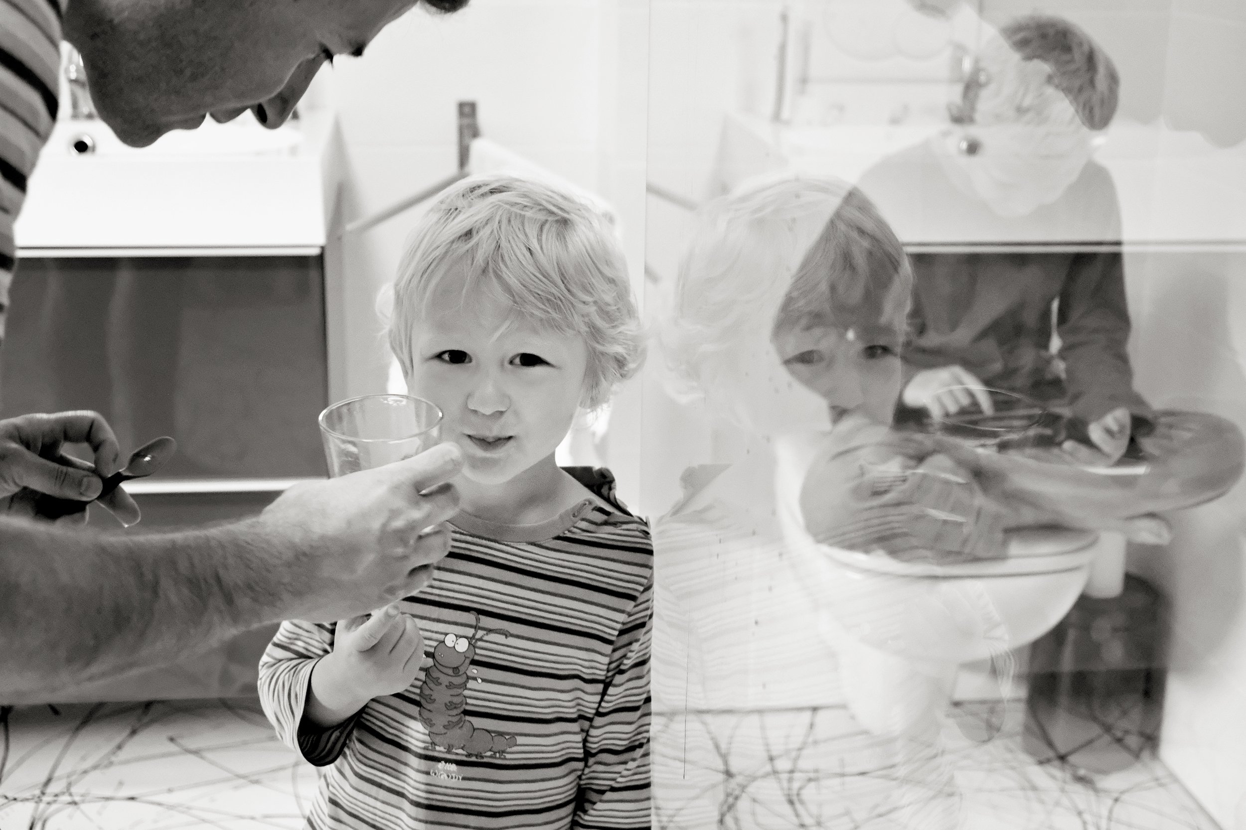 A young boy with blond hair holding a clear cup, smiling bashfully, standing behind a reflective glass barrier with a young girl behind it, both in a kitchen or indoor setting.