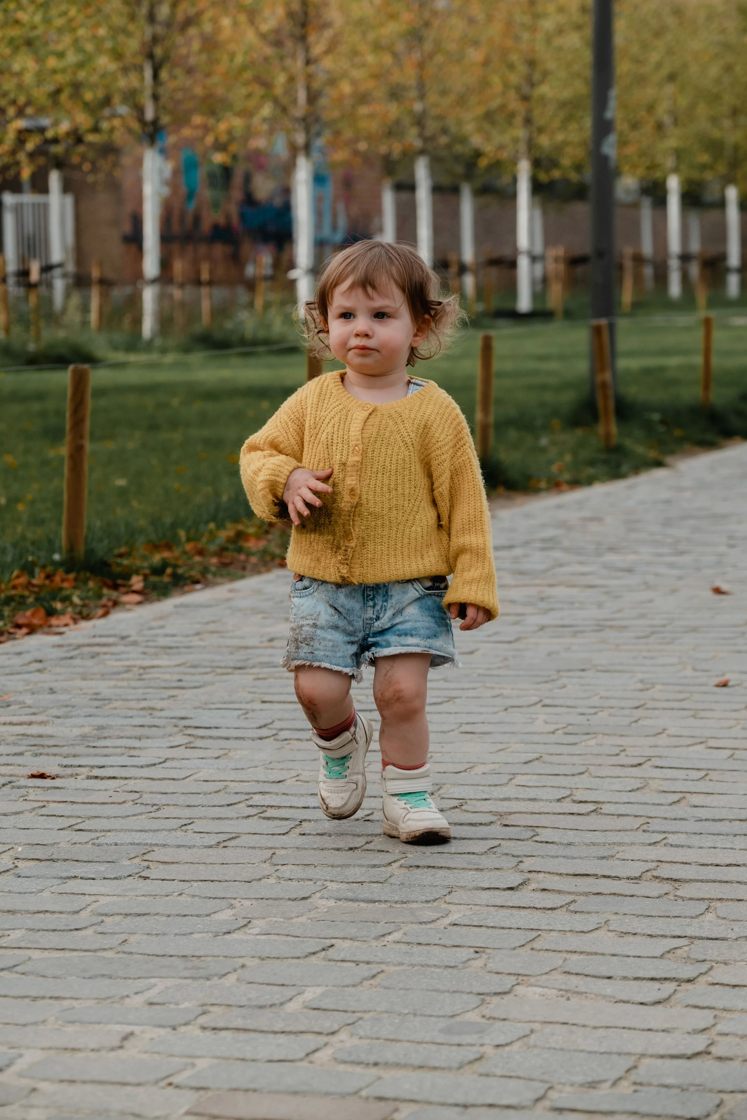 A young child with brown hair running on a paved path in a park during fall, wearing a yellow sweater, denim shorts, and sneakers.