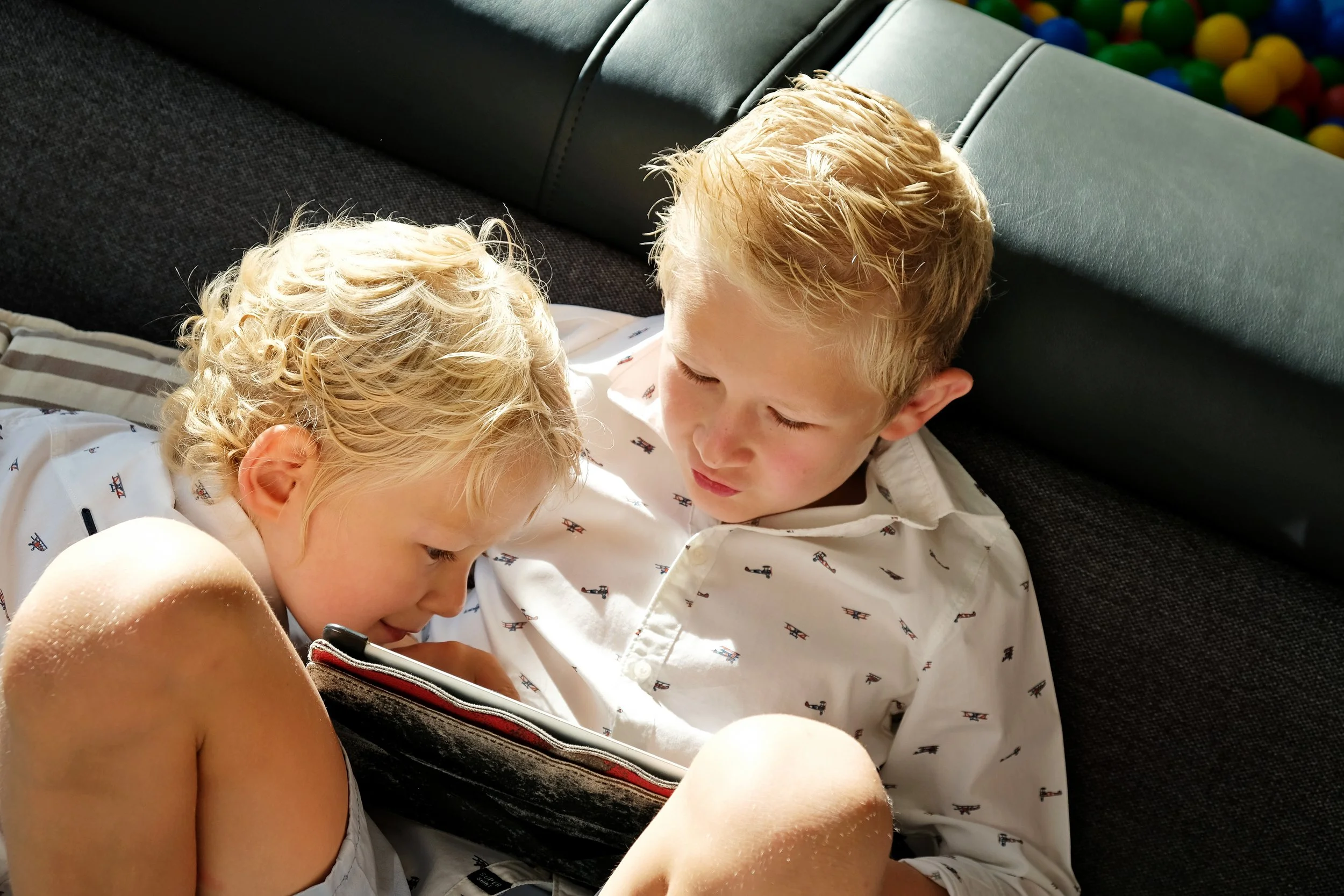 Two young children, a boy with blond hair and a girl with curly blond hair, sitting closely together on a black couch, looking at a phone in their hands. Behind them, there is a ball pit with colorful plastic balls.