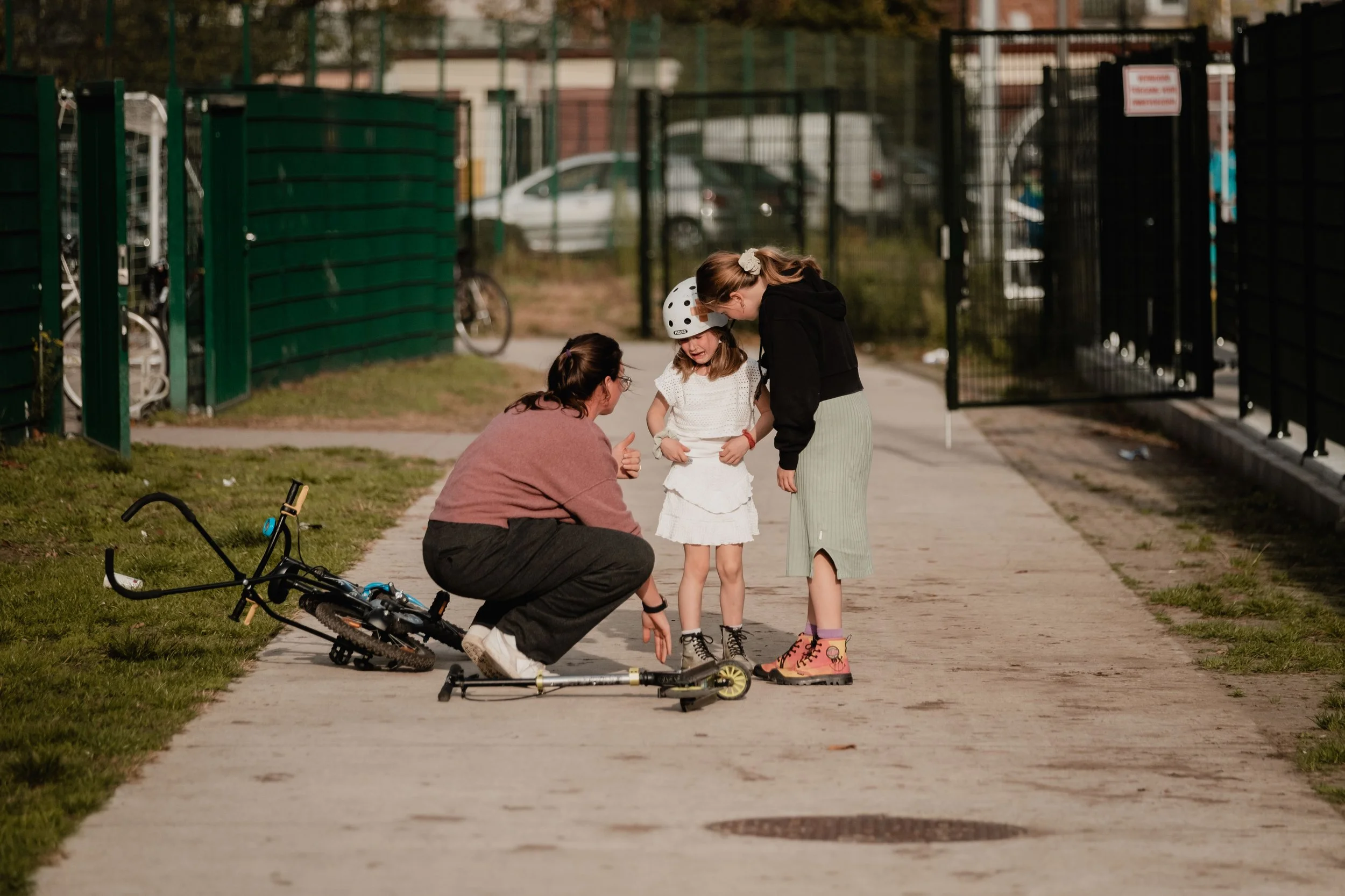 A woman kneeling beside a young girl wearing a helmet, both talking to another girl in rain boots, while a fallen bicycle lies on the sidewalk nearby.