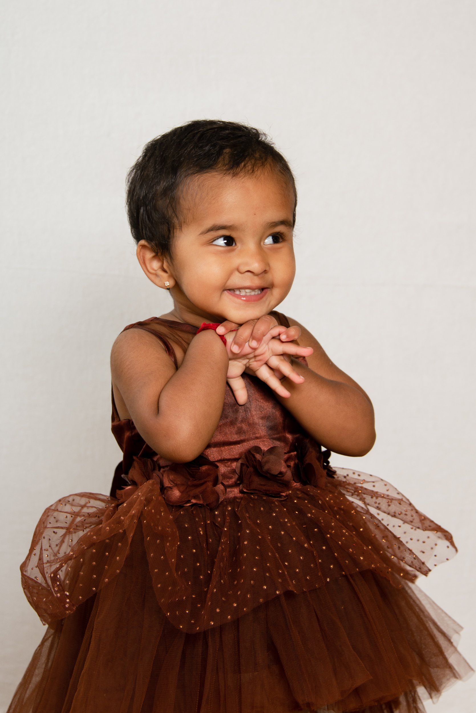 A young girl with short dark hair, wearing a brown and gold dress with a tulle skirt, smiling with hands clasped together, in front of a plain white background.